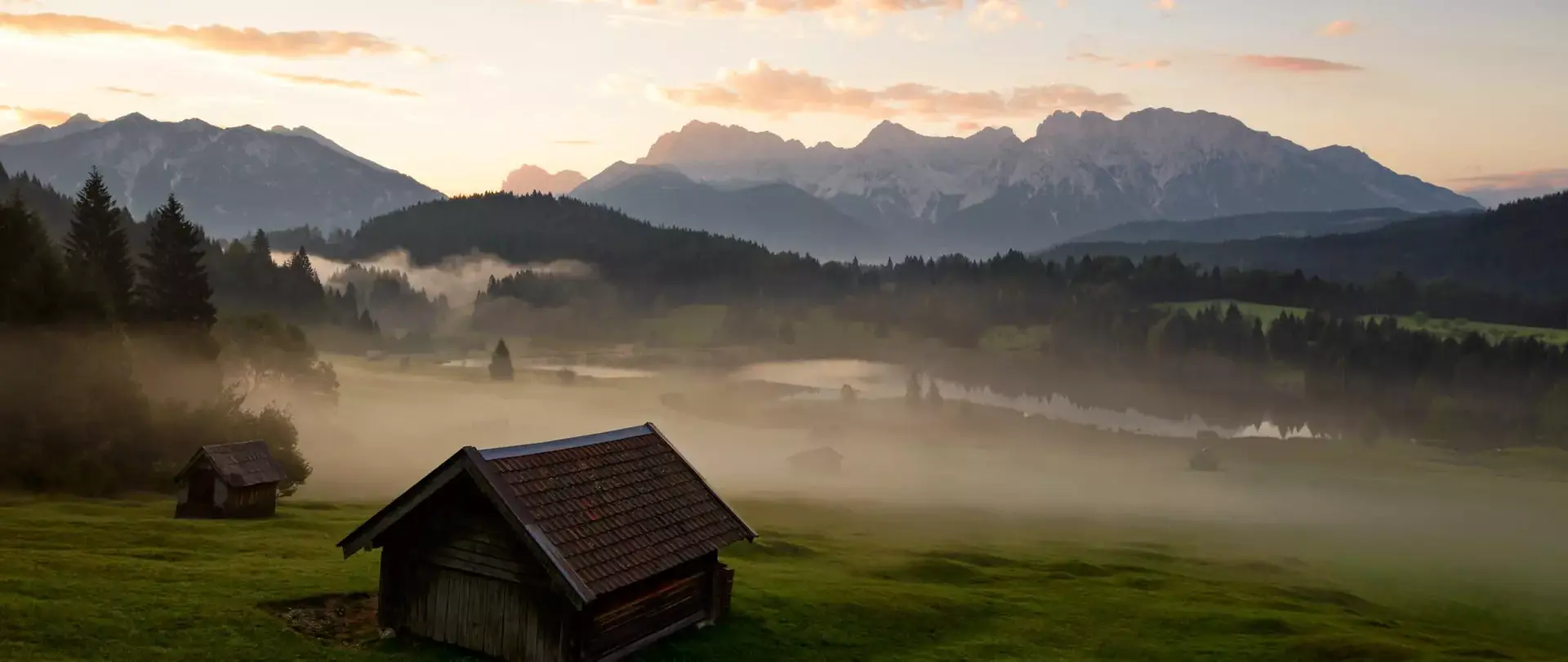 Morgenstimmung mit Nebel über grünen Wiesen und Holzhütten vor dem Karwendelgebirge in den Alpen