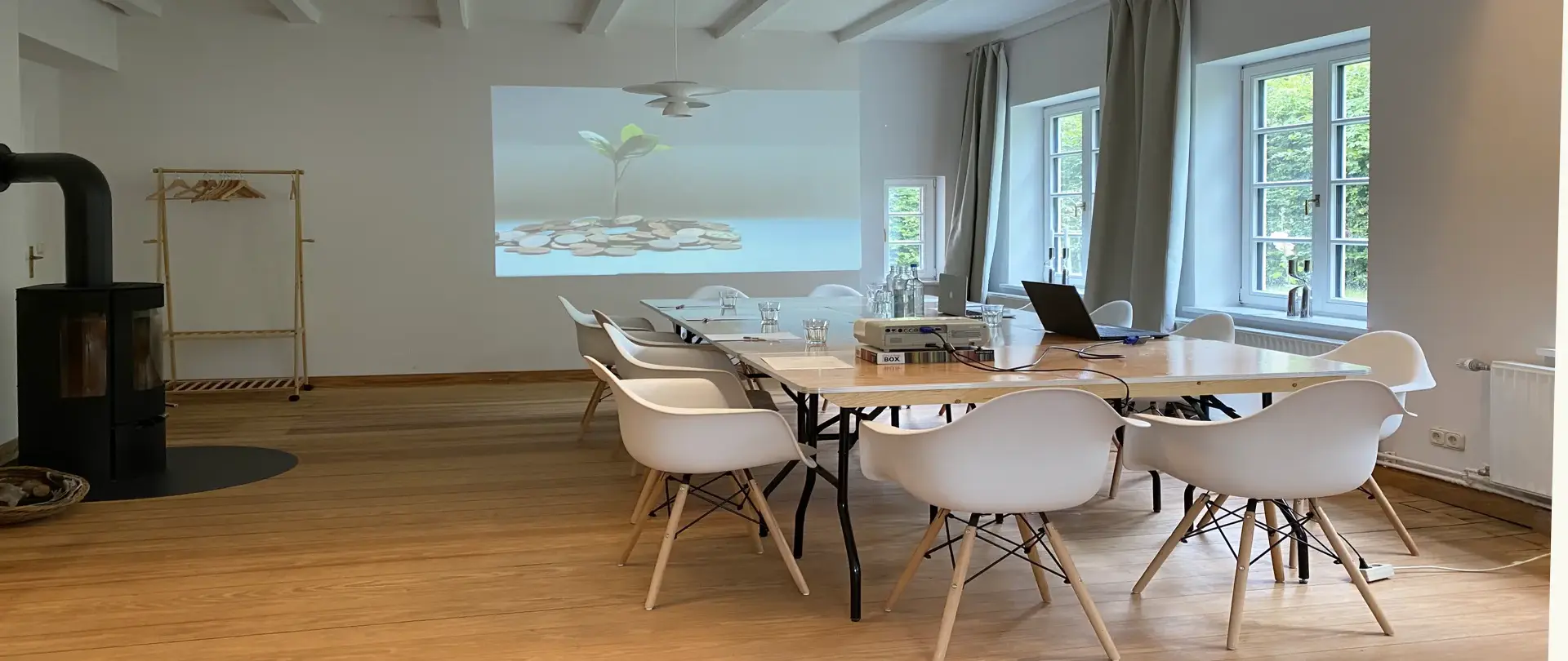Konferenzraum im Naturpark Aukrug mit Holzboden, weißen Stühlen, Holztisch, Laptop und Projektorwand mit Pflanze und Münzen