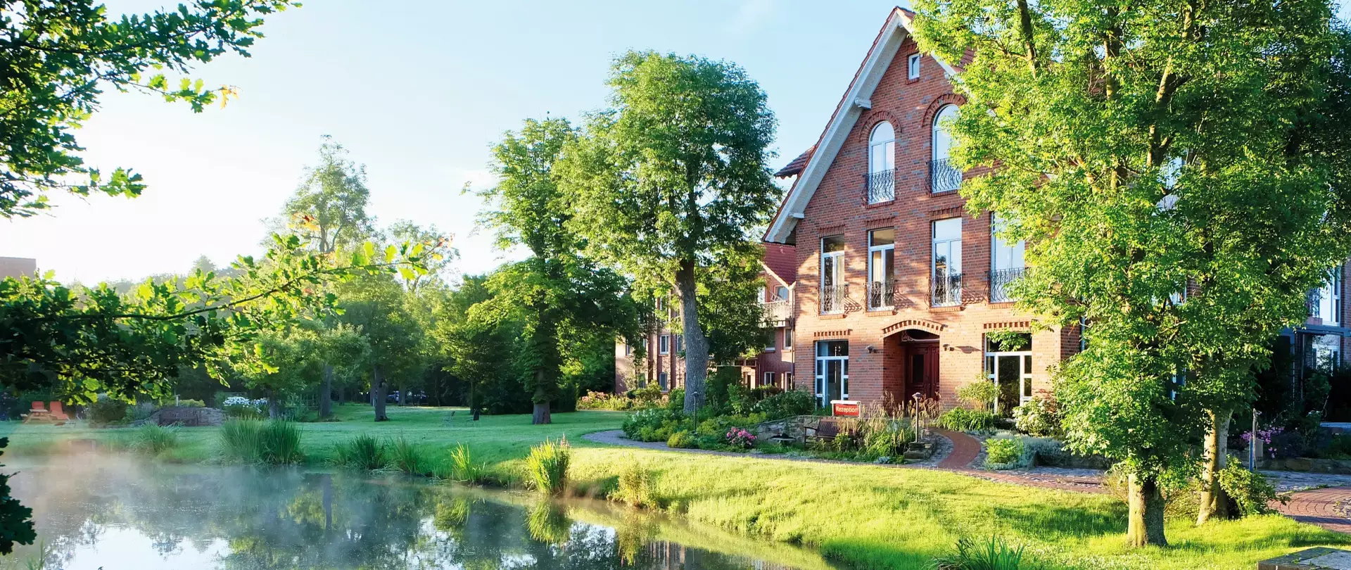 Backsteinhaus am Wasser mit Bäumen und grünem Ufer im Wattenmeer bei Sonnenlicht