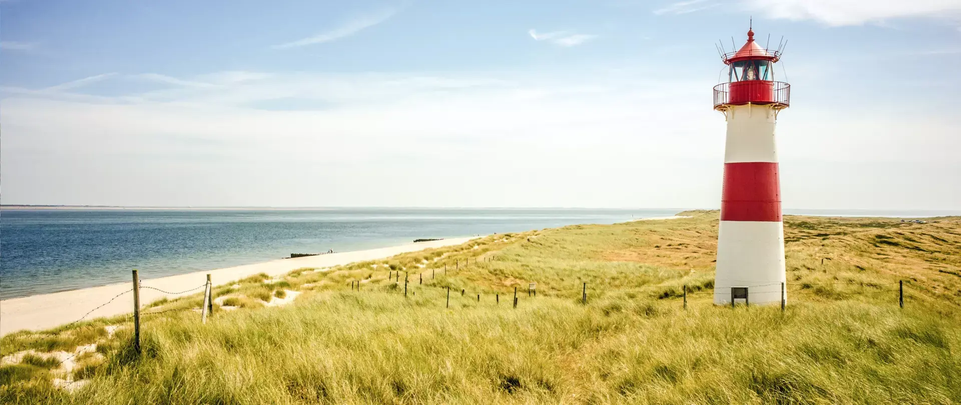 Leuchtturm mit roten Streifen an der Küste auf Sylt, umgeben von Dünengras und Sandstrand unter blauem Himmel