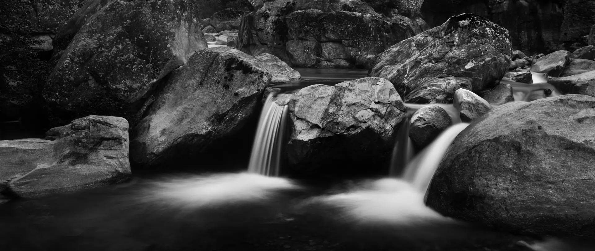 Schwarzweißaufnahme eines leise fließenden Bachs mit Wasserfällen zwischen großen Felsen, die Ruhe und reduzierte Kontraste der Fine Art Fotografie zeigen