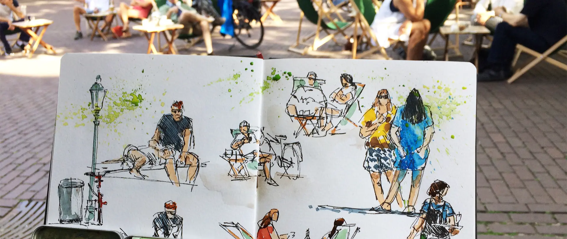 Skizzenbuch mit Aquarellzeichnungen von Menschen auf Stühlen in einem belebten Außenbereich bei Sonnenlicht