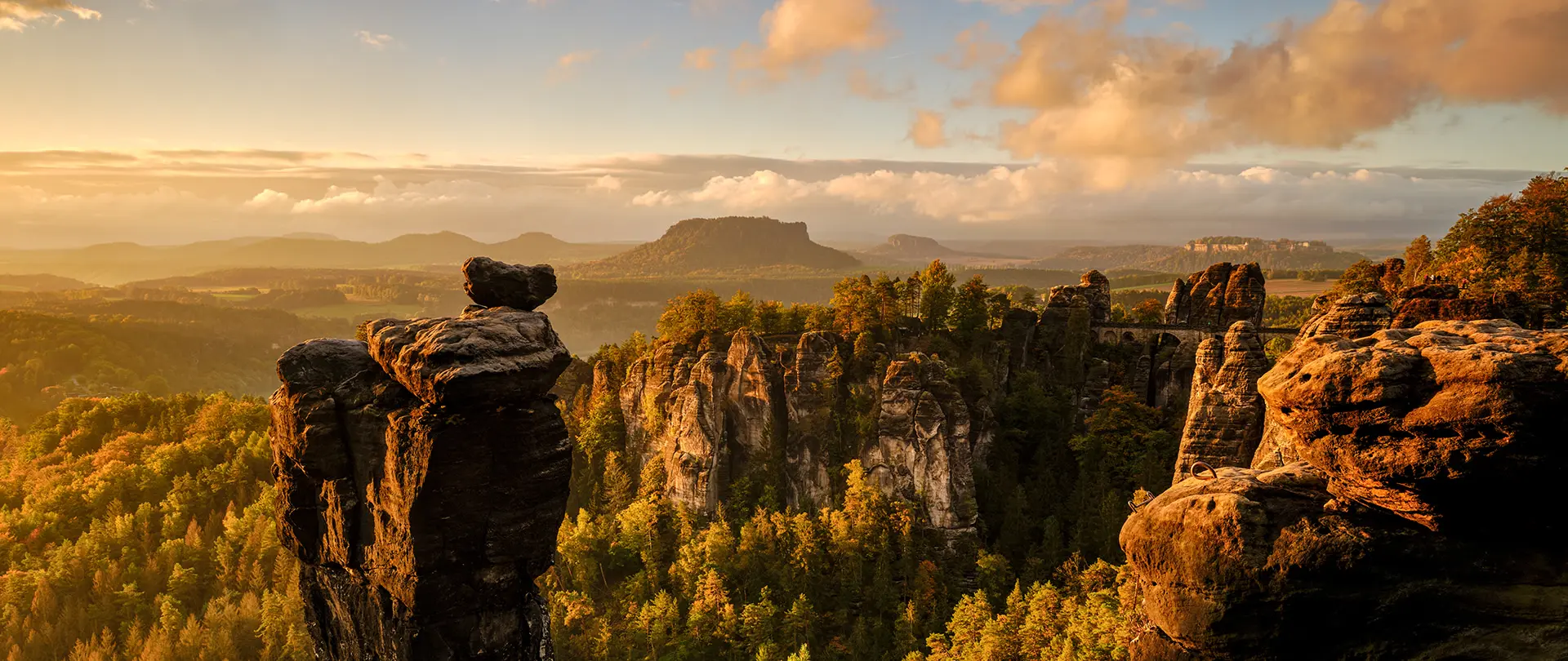 Felsformationen und bewaldete Landschaft der Sächsischen Schweiz im warmen Licht des Sonnenuntergangs mit Blick auf Tafelberge im Hintergrund