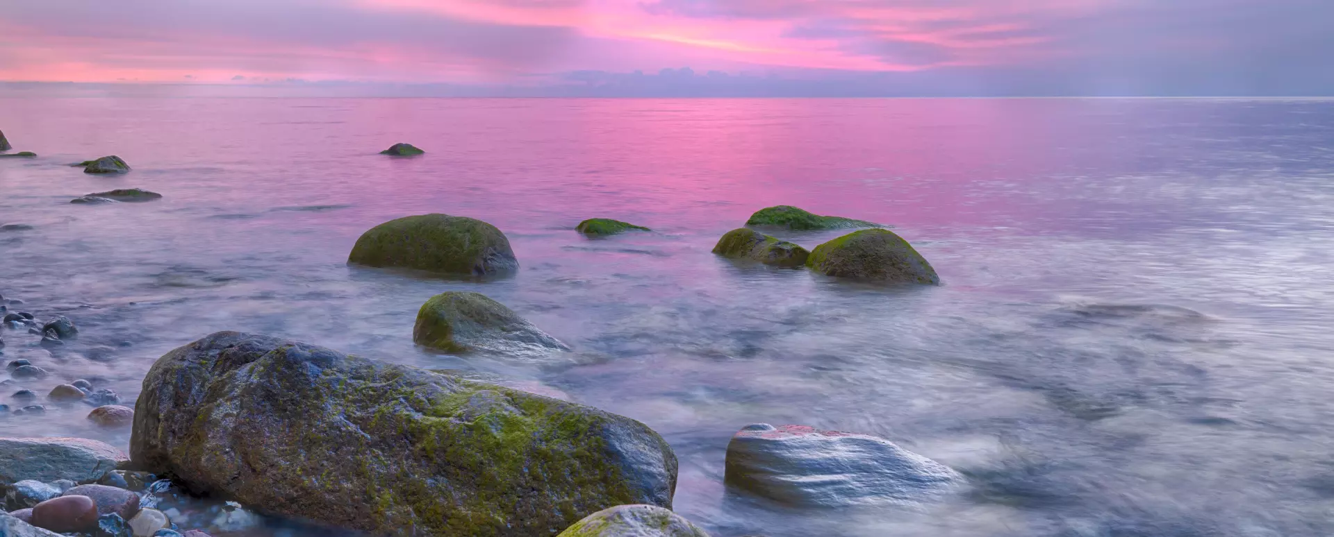 Blick auf eine ruhige Ostseeküste mit moosbedeckten Felsen im Wasser bei Sonnenuntergang auf Rügen, Deutschland. Der Himmel ist in Rosa- und Lilatönen gefärbt.