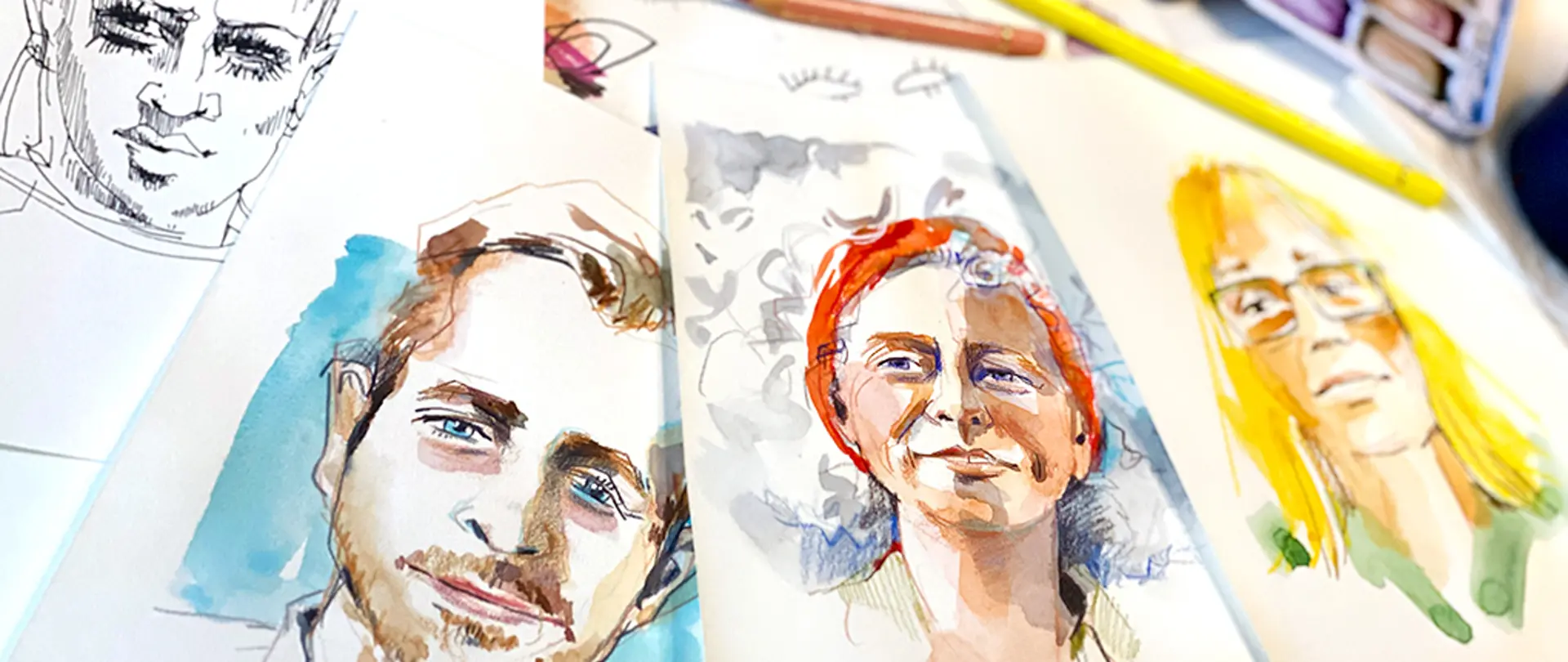 Mehrere handgezeichnete und aquarellierte Portraits auf Papier, die entspannte Zeichenatmosphäre eines Wochenendporträtkurses mit Stiften und Farben zeigen