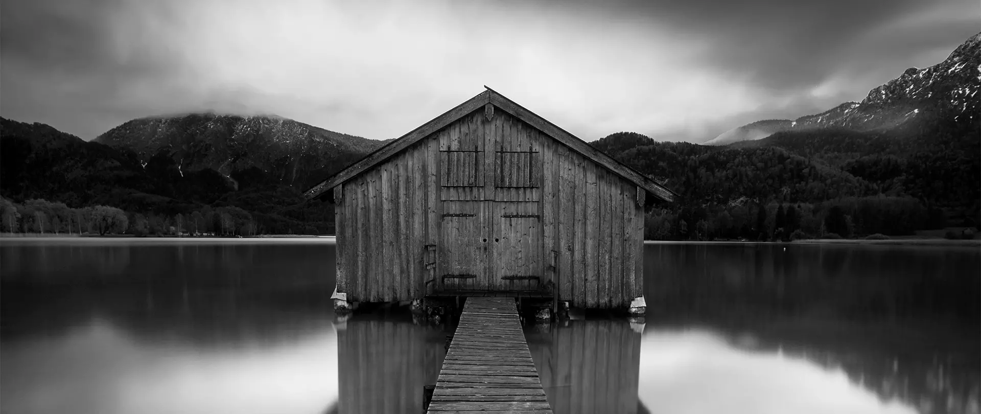 Schwarz-weiß-Fotografie eines hölzernen Bootshauses auf einem See, umgeben von Bergen und bewölktem Himmel erstellt von artistravel Dozenten Thomas Bichler