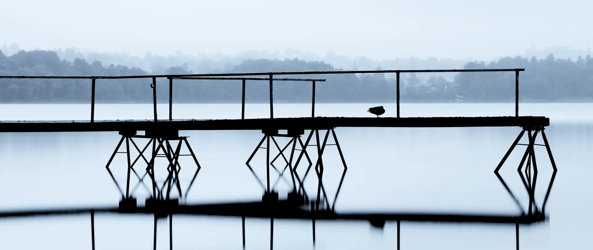Steg mit Geländer über ruhigem Bergsee, Reflexion im Wasser und neblige Berglandschaft im Hintergrund, minimalistische Naturfotografie
