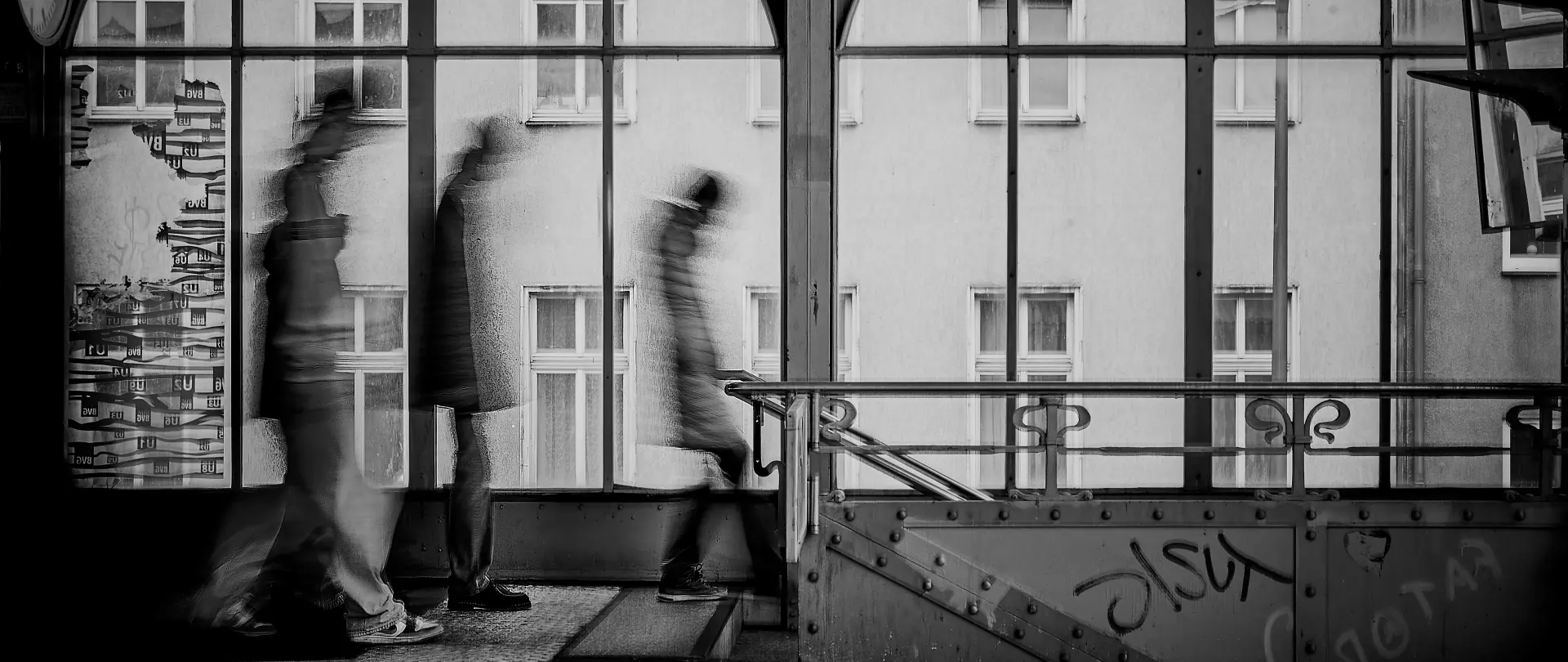 Schwarz-weiß-Foto von verschwommenen Menschen in Bewegung vor urbaner Glasfassade mit barocker Treppengeländerzeichnung und Berliner Stadtarchitektur im Hintergrund