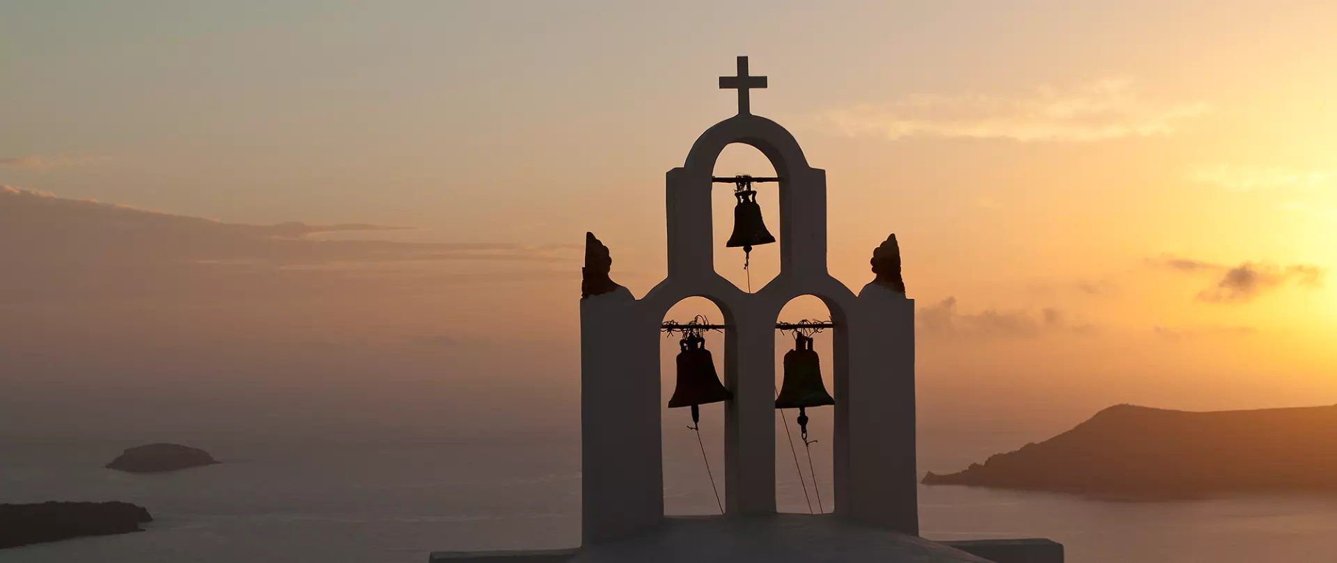 Kirchturmglocken einer Kapelle auf Santorin vor orangefarbenem Sonnenuntergang über der Ägäis mit Inseln am Horizont