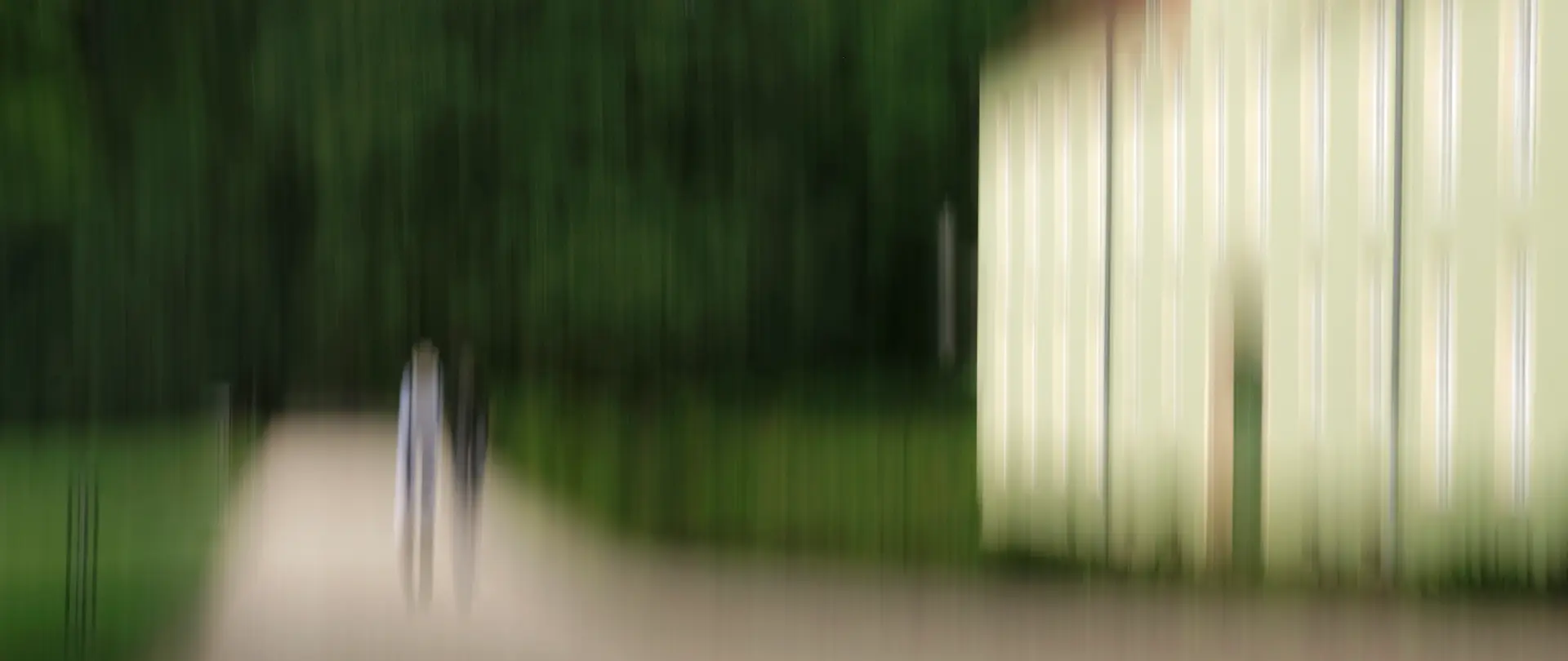 Zwei Personen spazieren auf einem Weg neben einem historisch wirkenden Gebäude in einem grünen Park, unscharf und künstlerisch verwischt