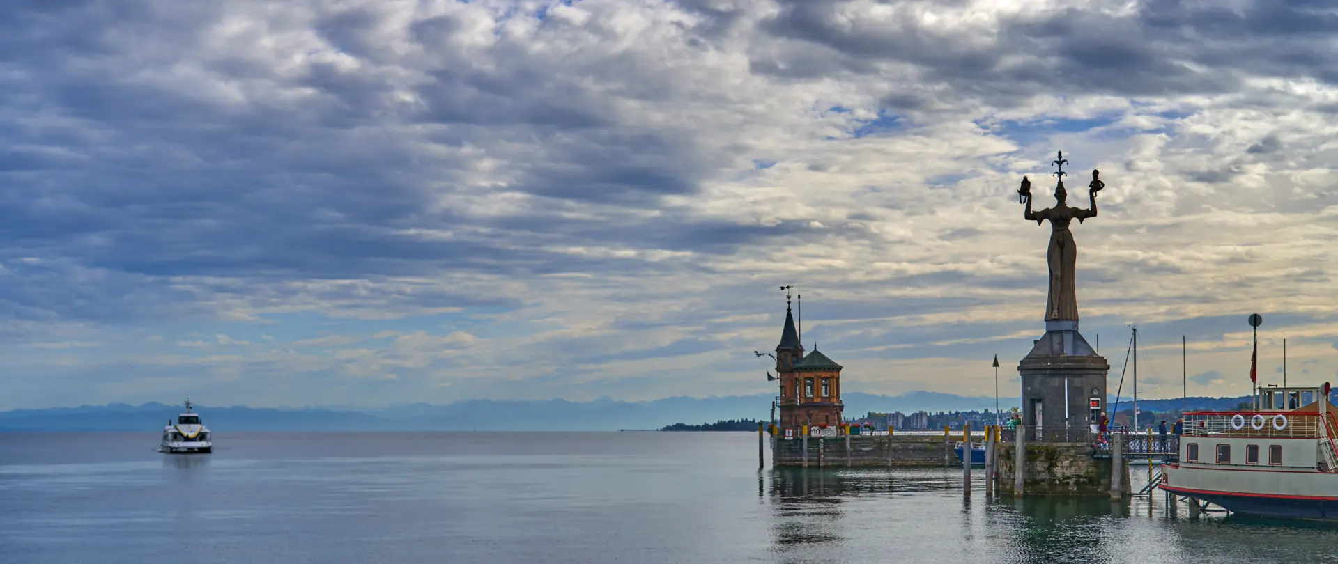 Bodensee mit Löwenbrunnen, historischen Bauten und Boot, ruhige Wasseroberfläche unter bewölktem Himmel als Kulisse für visuelle Geschichten