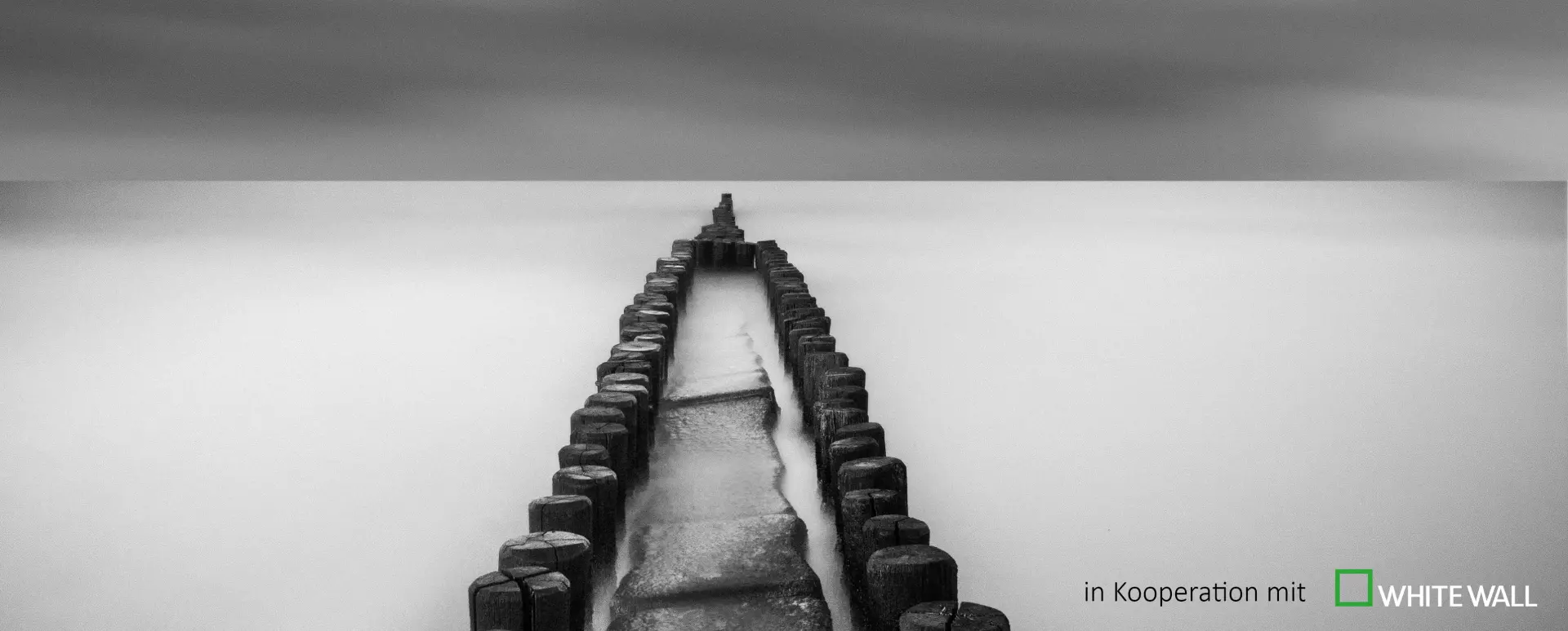 Schwarz-weiß-Foto eines minimalistischen, symmetrischen Holzstegs, der in ruhigem Wasser mit nebligem Hintergrund ins Unendliche führt