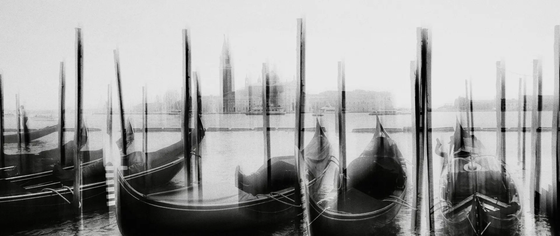 Schwarz-weiß Aufnahme von venezianischen Gondeln vor der Stadt mit verschwommenem Effekt, der die lebendige und vielfältige Atmosphäre Venedigs vermittelt
