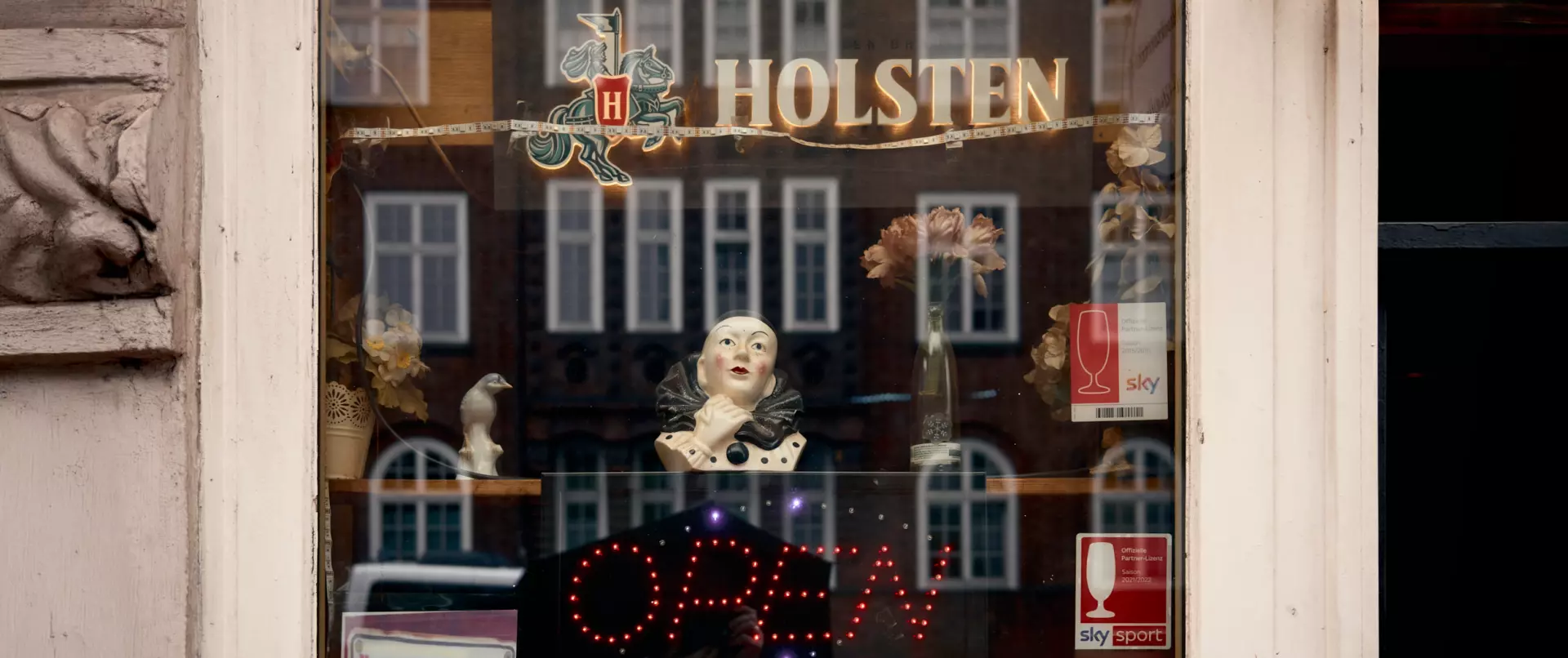 Schaufenster mit Holsten-Logo, Künstlerskulpturen und offen leuchtendem "OPEN"-Schild vor Spiegelung von Stadthäusern, urbaner Motivsuche abseits typischer Klischees