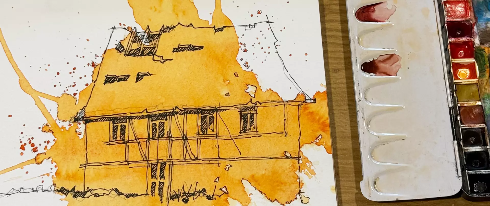 Aquarellskizze eines Fachwerkhauses mit orangener Farbfläche und schwarzen Fineliner-Linien, daneben ein geöffneter Aquarellkasten mit genutzten Farbnäpfchen.