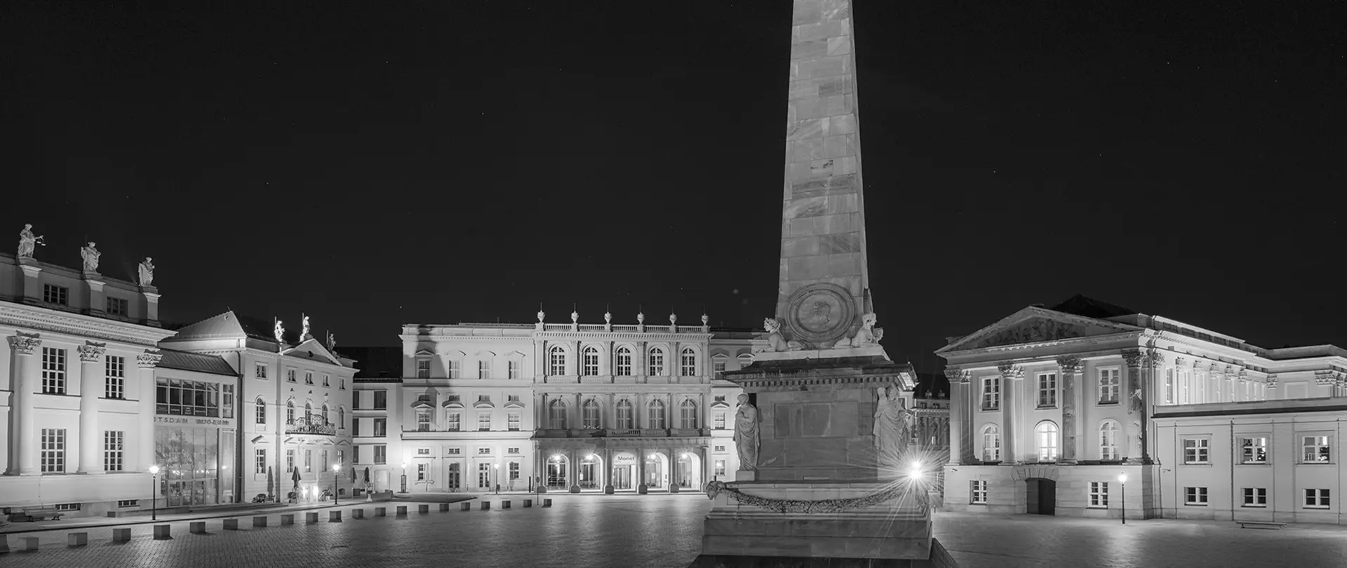 Monochrome Aufnahme des Alten Marktes in Potsdam mit beleuchteten historischen Gebäuden und einem Obelisken im Vordergrund bei Nacht