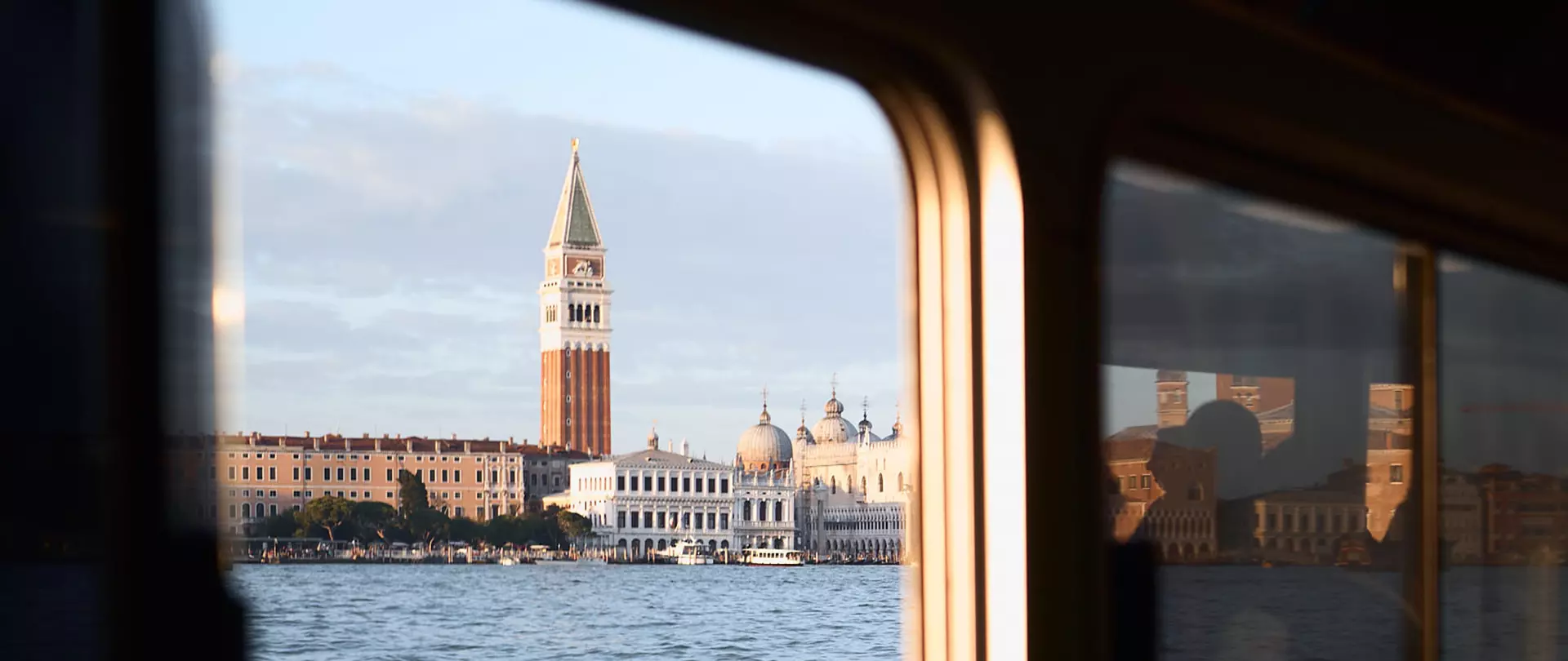 Blick auf den Glockenturm in Venedig, aus der Perspektive eines Bootes. Die Fenster des Bootes sind am Bildrand zu erkennen sowie Spiegelungen von Personen.