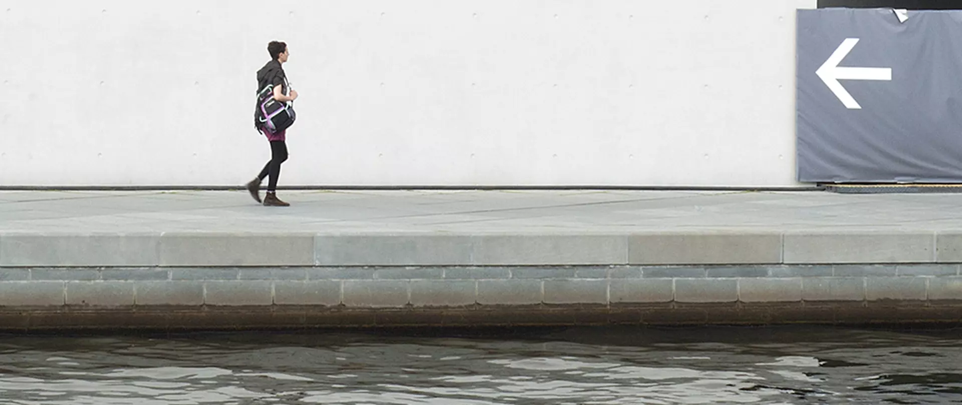 Frau mit Rucksack geht an einem Wasserkanal entlang, weiße Wand mit großem Pfeil nach links fotografiert von artistravel Dozentin Jutta Engelage Fotografie