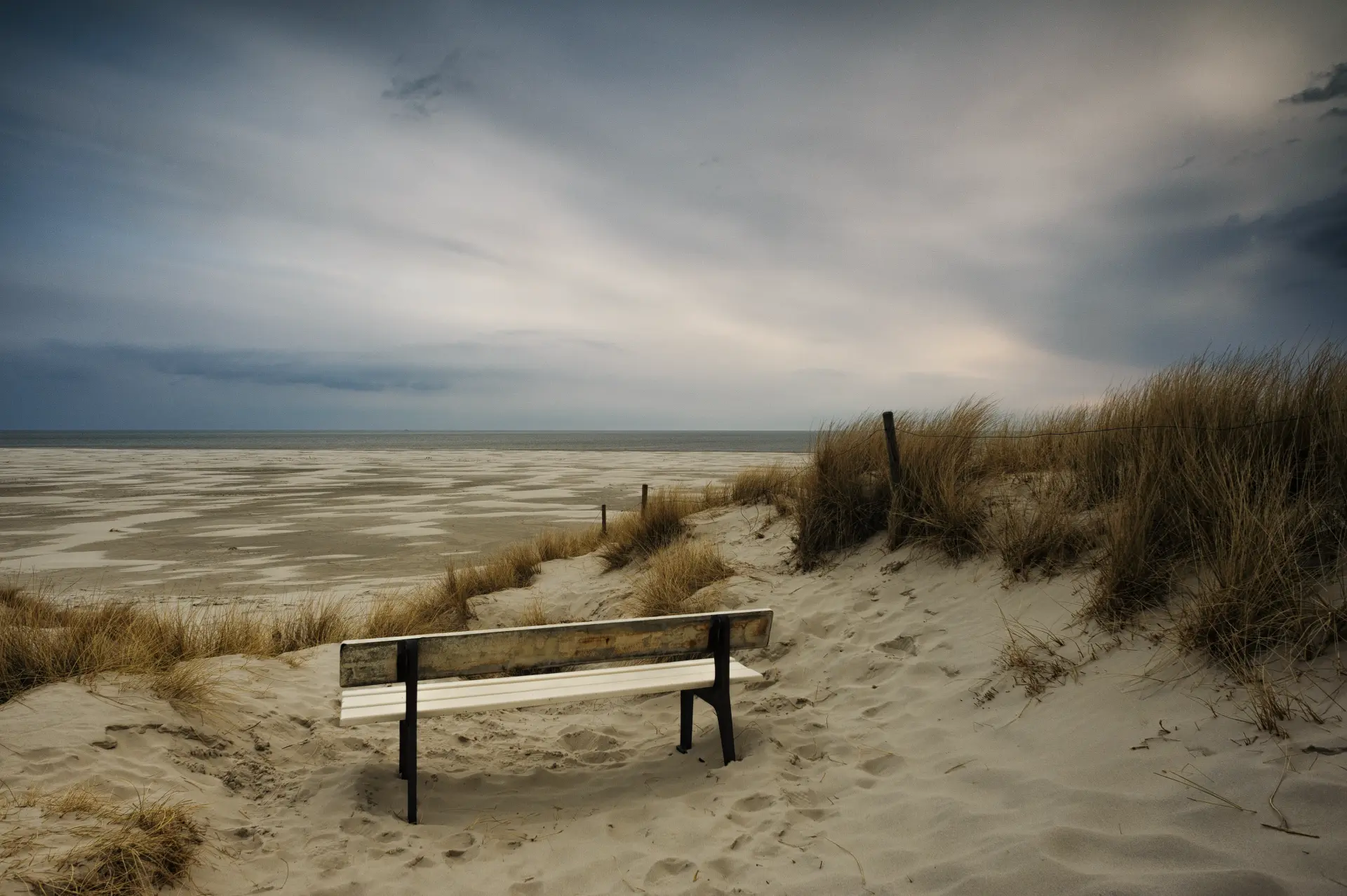 Leere Holzbank in den Dünen der nordfriesischen Insel Amrum mit Blick auf den ruhigen, weiten Nordseestrand unter bewölktem Himmel