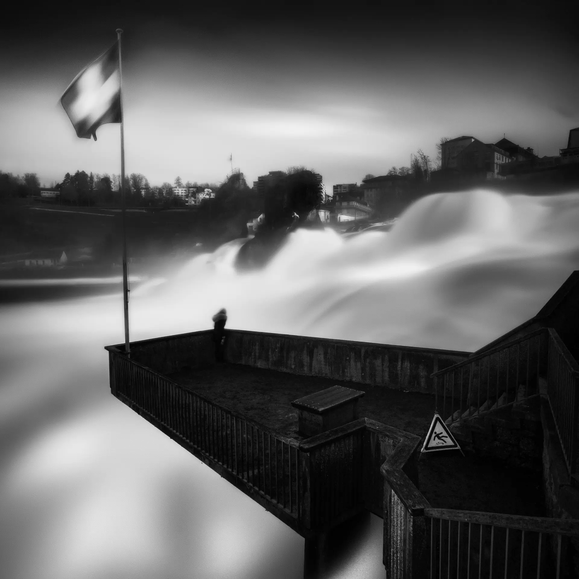 Schwarz-weiß Fine Art Fotografie am Bodensee mit fließendem Wasserfall, Schweizer Flagge und stimmungsvoller Landschaft im Hintergrund