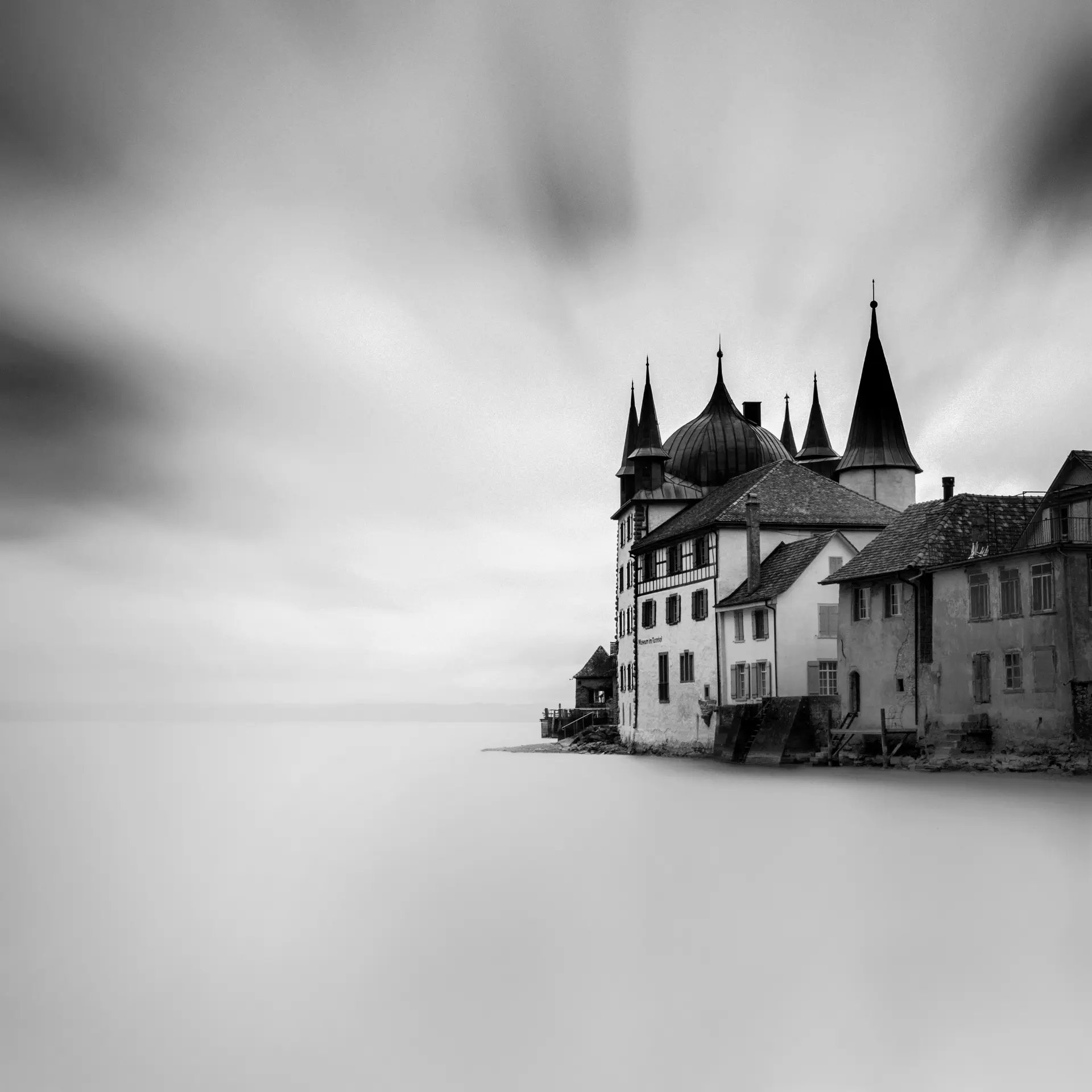 Schwarz-Weiß-Fine-Art-Fotografie eines historischen Gebäudes mit Türmen am ruhigen westlichen Ufer des Bodensees unter bewölktem Himmel