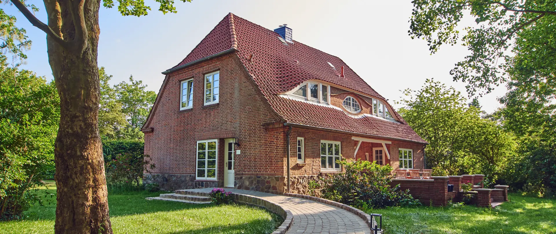 Backsteinhaus mit rotem Satteldach und gepflastertem Weg, umgeben von Bäumen und grüner Wiese im Naturpark Aukrug
