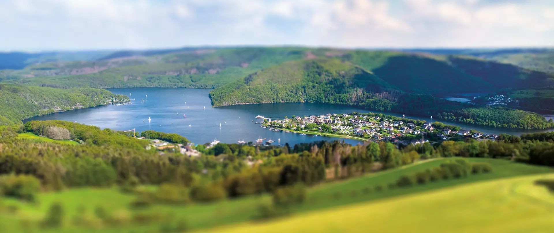 Blick auf einen See in der Eifel mit umliegenden Wäldern, Hügeln und einem kleinen Dorf am Wasserufer unter einem bewölkten Himmel