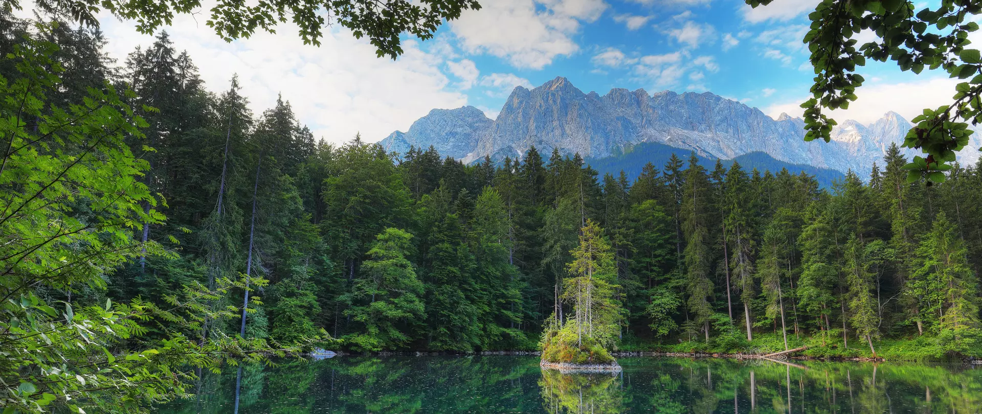 Bergsee in der Karwendelregion umgeben von dichtem Wald und Bergen unter blauem Himmel mit Wolken
