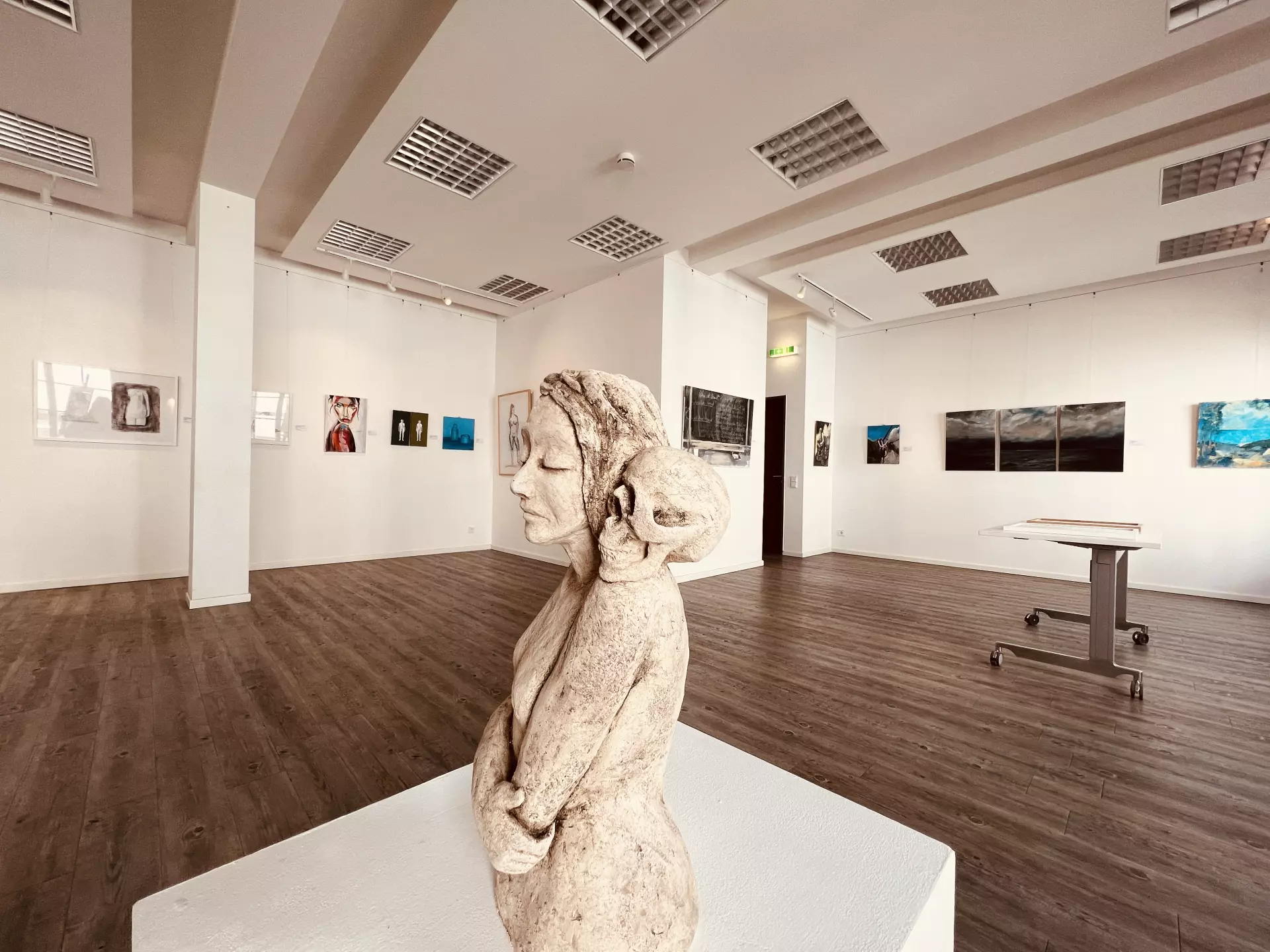 Skulptur einer Frau mit geschlossenen Augen in einer modernen Galerie in der Eifel, umgeben von Gemälden an weißen Wänden und hellem Holzfußboden