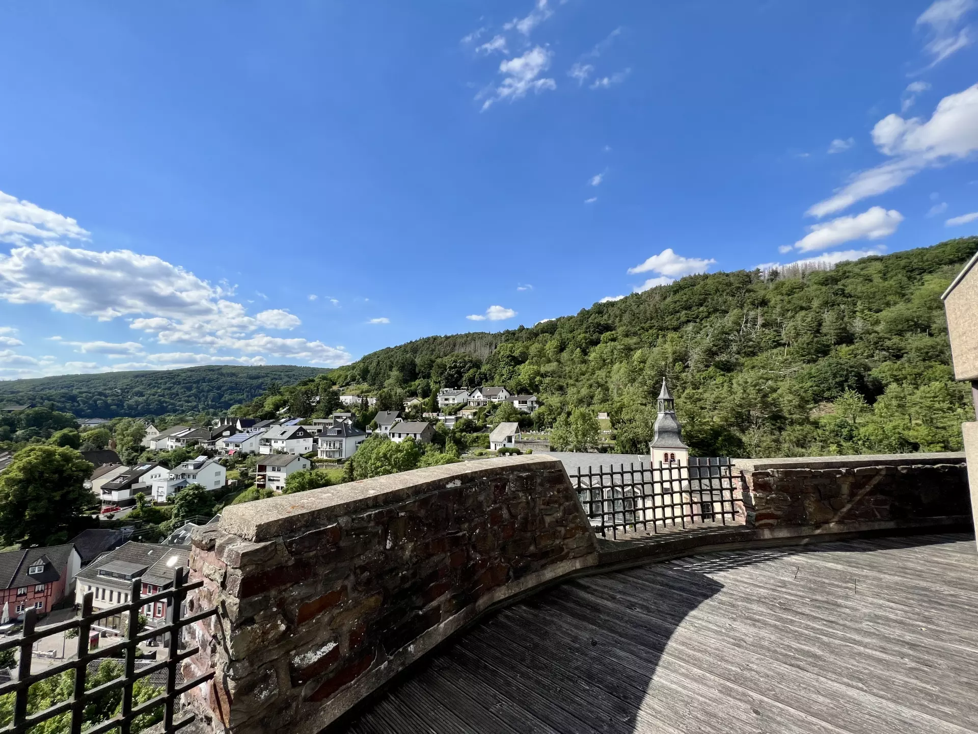 Aussichtsplattform mit Steinmauer über einem Eifeldorf, grünen Hügeln und blauem Himmel mit vereinzelten Wolken