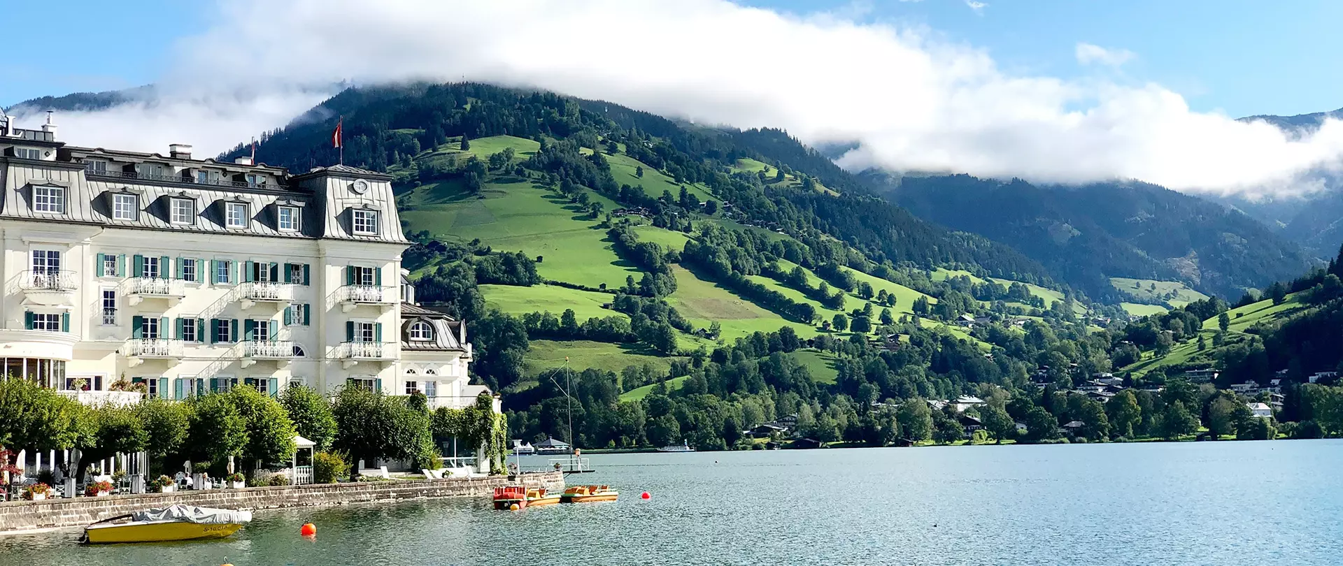 Hotelgebäude am Ufer eines Sees mit grünen bewaldeten Bergen und Wolken im Hintergrund in Zell am See