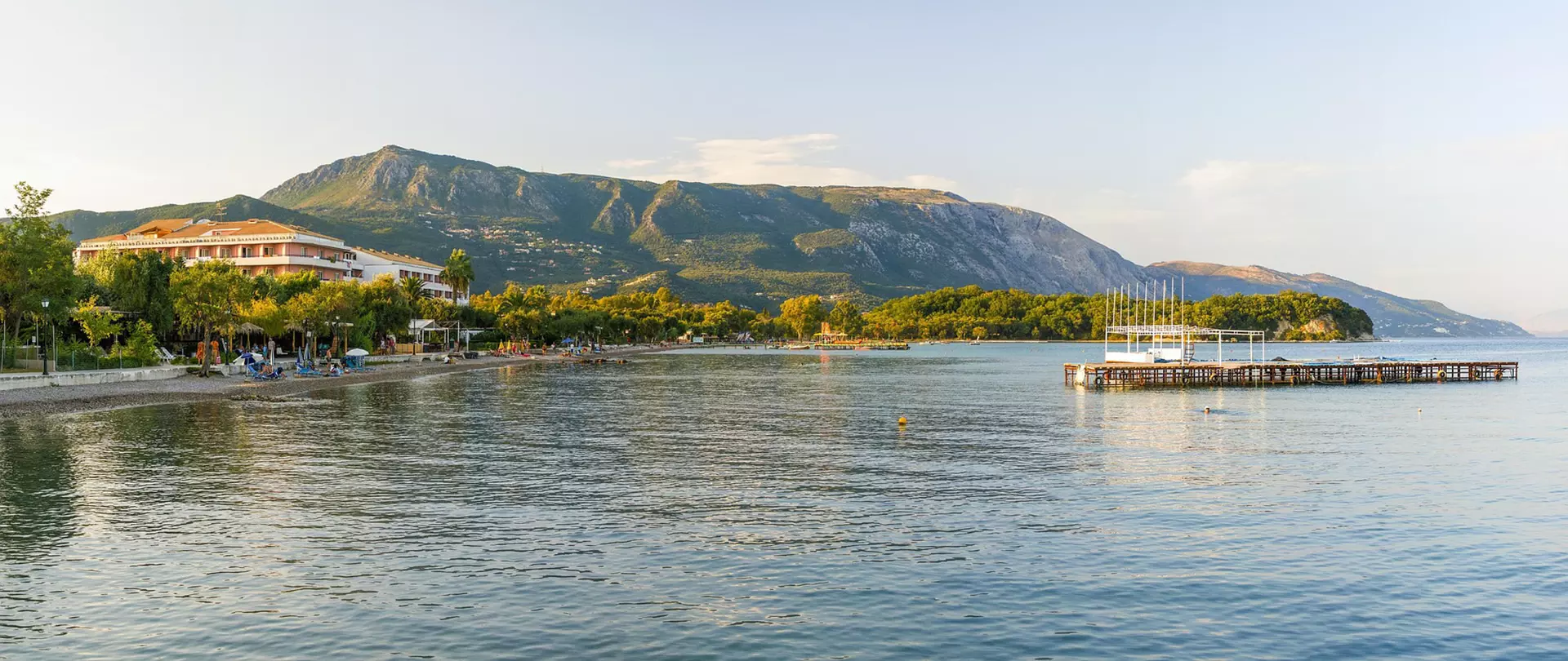 Strand und Promenade von Agios Gordios auf Korfu mit Bergkulisse und schwimmendem Steg im ruhigen Meer bei klarem Himmel