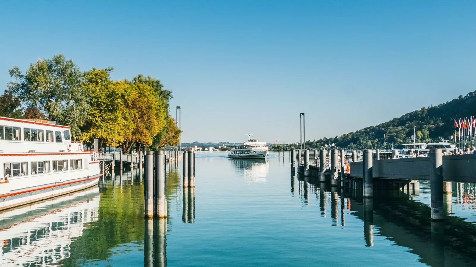 Christiane Setz, Bregenz Tourismus Hafen in Bregenz mit Passagierschiff, Stegen, Bäumen und Hügel im Hintergrund bei klarem Himmel