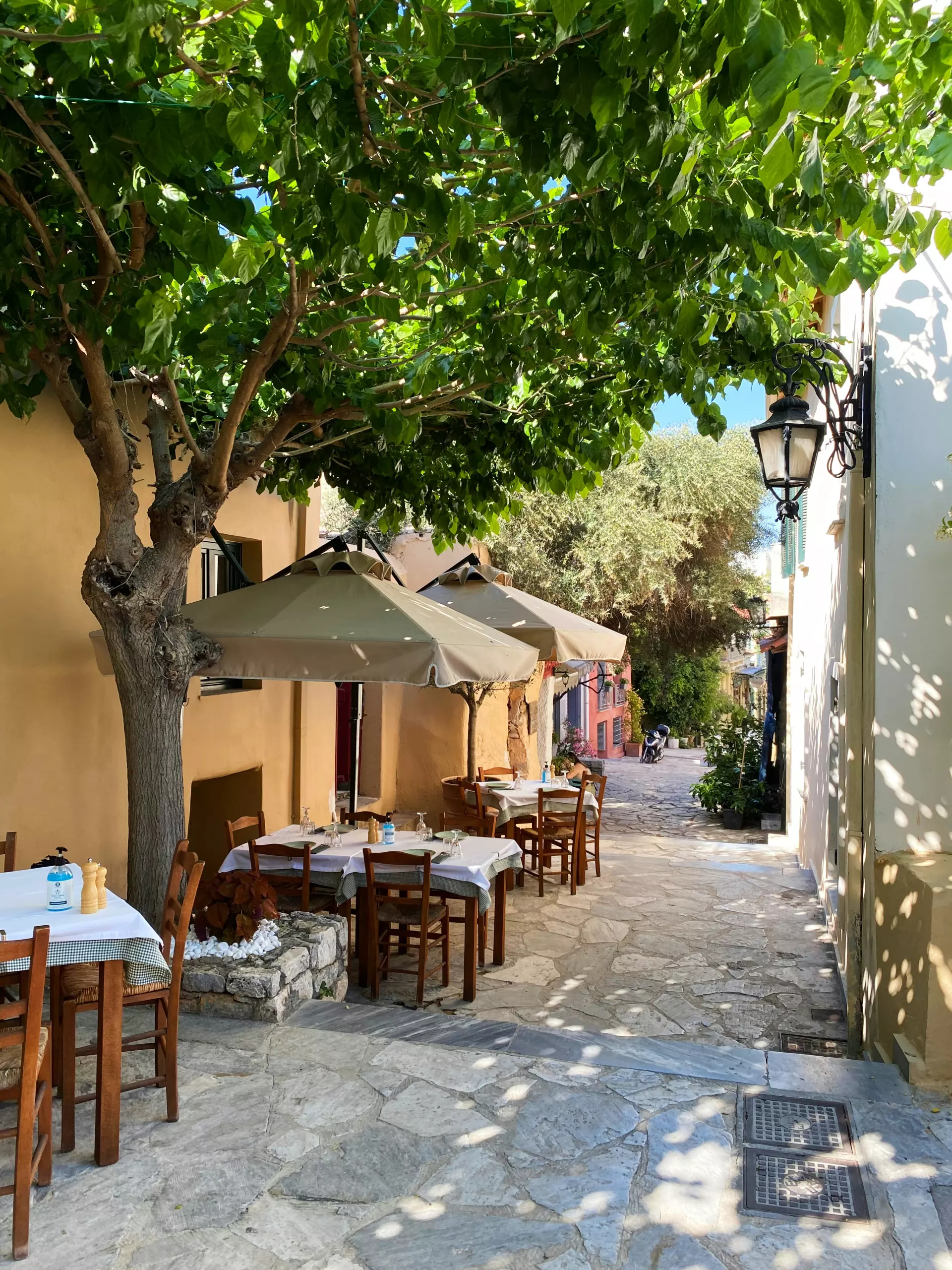 Straße in Athen mit Außensitzplätzen eines Cafés, Tischen unter Sonnenschirmen und grünen Bäumen an sonnigem Tag