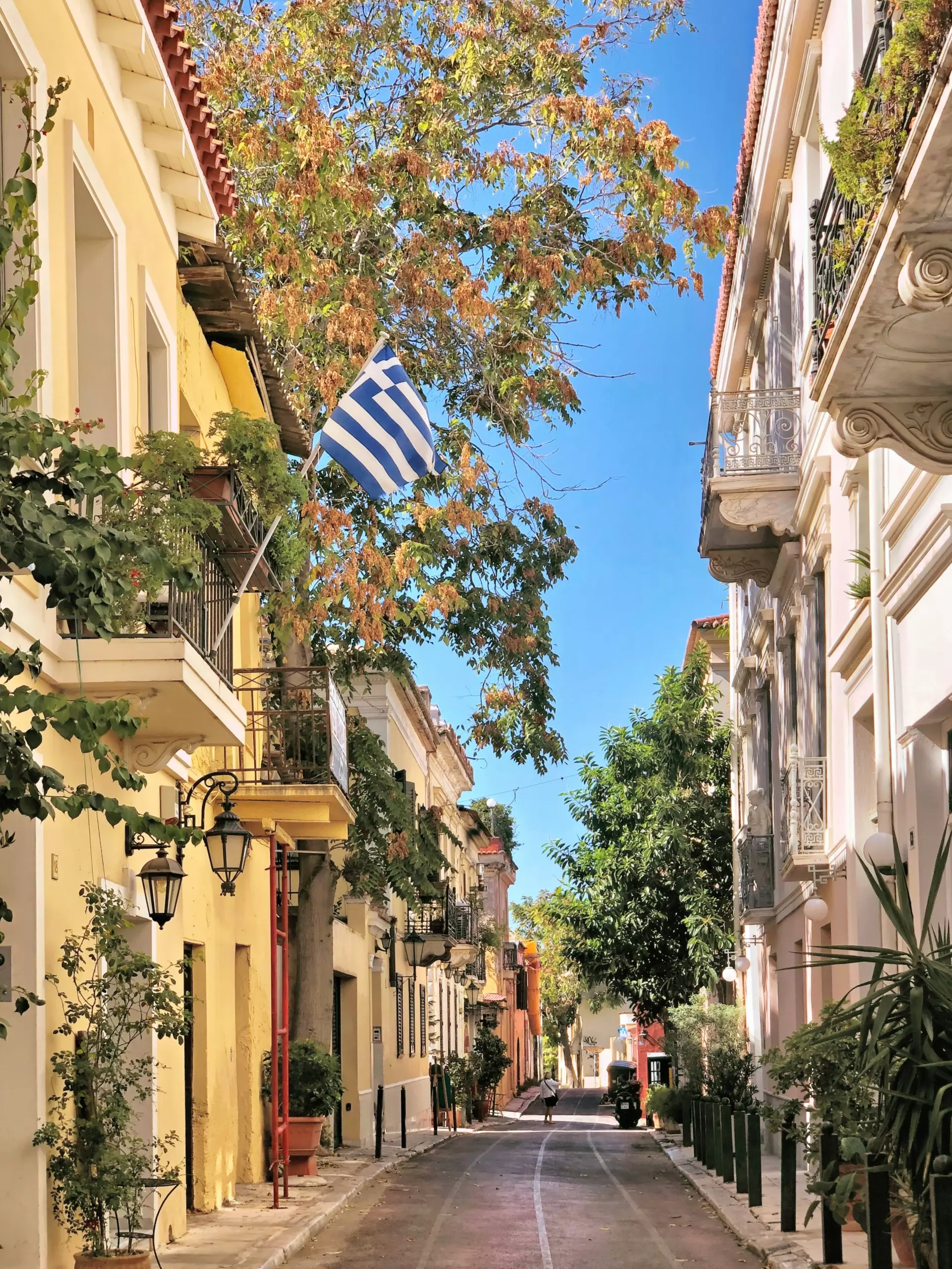 Schmale Straße in Athen mit griechischer Flagge, bunten Häusern, Balkonen und Bäumen unter klarem blauem Himmel
