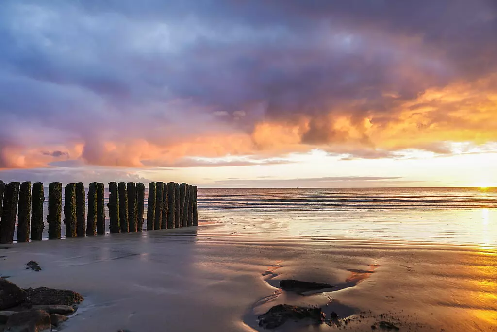 Sonnenuntergang über dem Wattenmeer mit Holzpfählen am Strand von Borkum und reflektierendem Wasser im feinen Sand