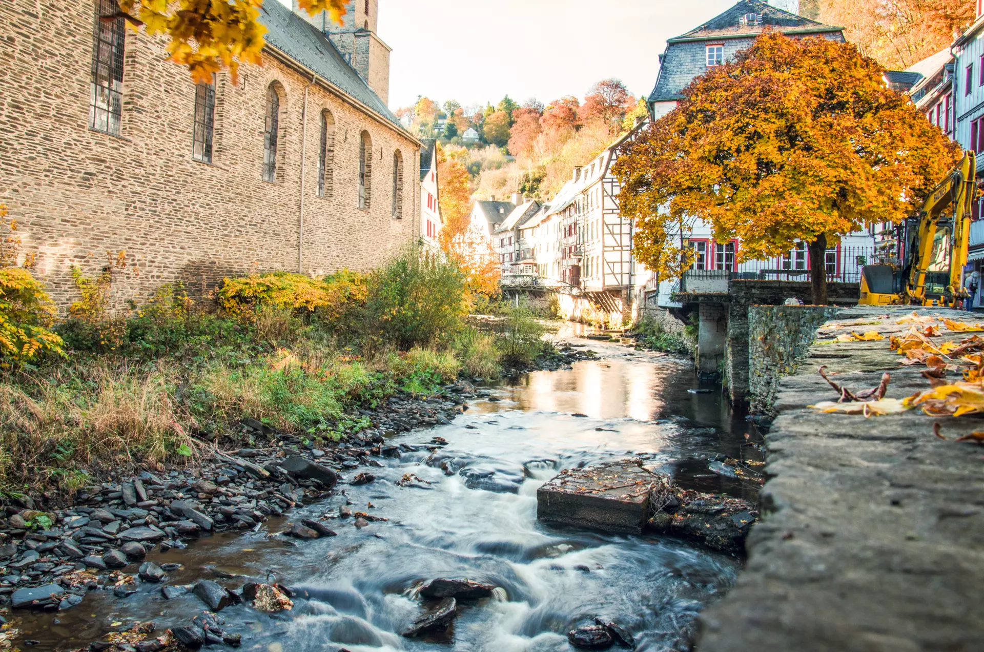 Herbstliche Eifellandschaft mit Bach, historischen Fachwerkhäusern, Kirche und buntem Laub an Bäumen im fließenden Wasser entlang eines Steinwegs