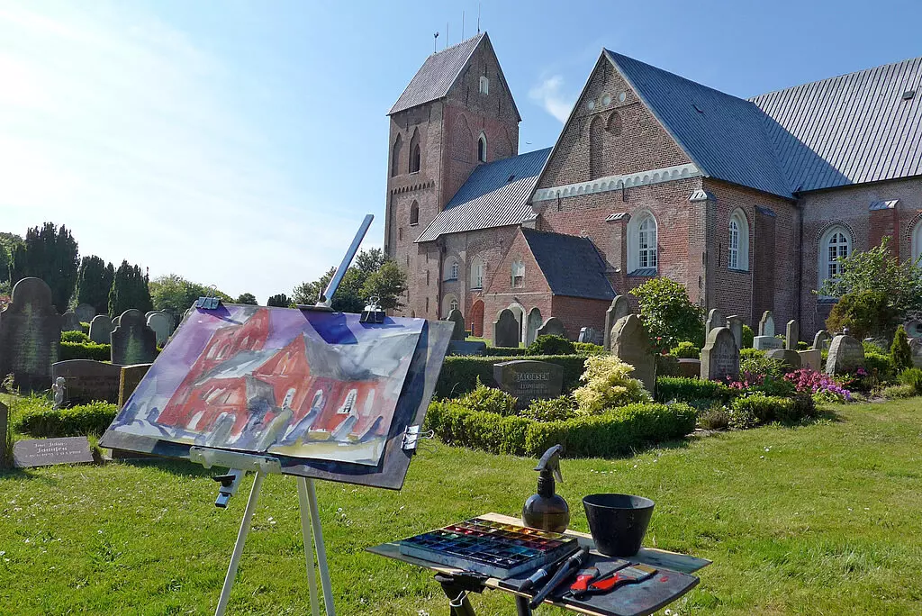 Kurseinblick Aquarellbild einer Kirche in Föhr auf Staffelei vor der echten Kirche mit Friedhof und Malutensilien bei sonnigem Wetter