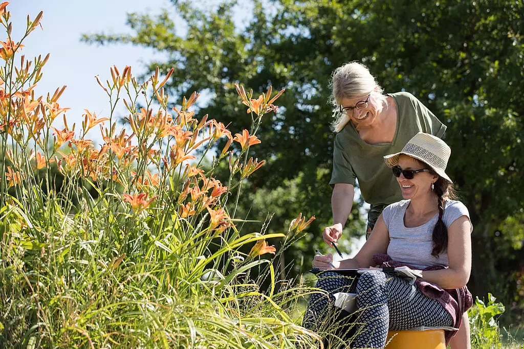 Zwei Frauen, eine im Rollstuhl mit Sonnenhut und die andere stehend, genießen gemeinsam eine sonnige Gartenszenerie mit blühenden orangen Taglilien in der Moselregion