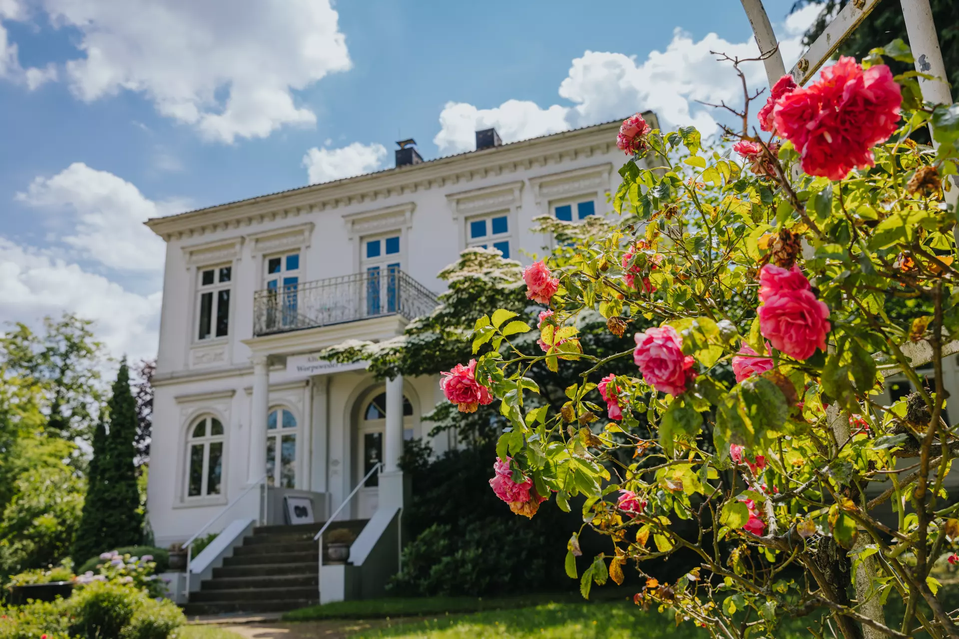 Weißes historisches Haus in Worpswede mit blühenden roten Rosen im Vordergrund und blauem Himmel mit Wolken