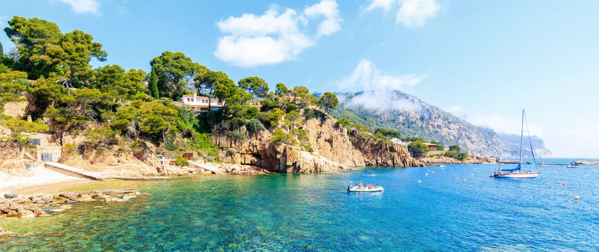 Küste von Mallorca mit kristallklarem Wasser, Segelboot, Motorboot und bewaldeten Felsen unter blauem Himmel mit wenigen Wolken