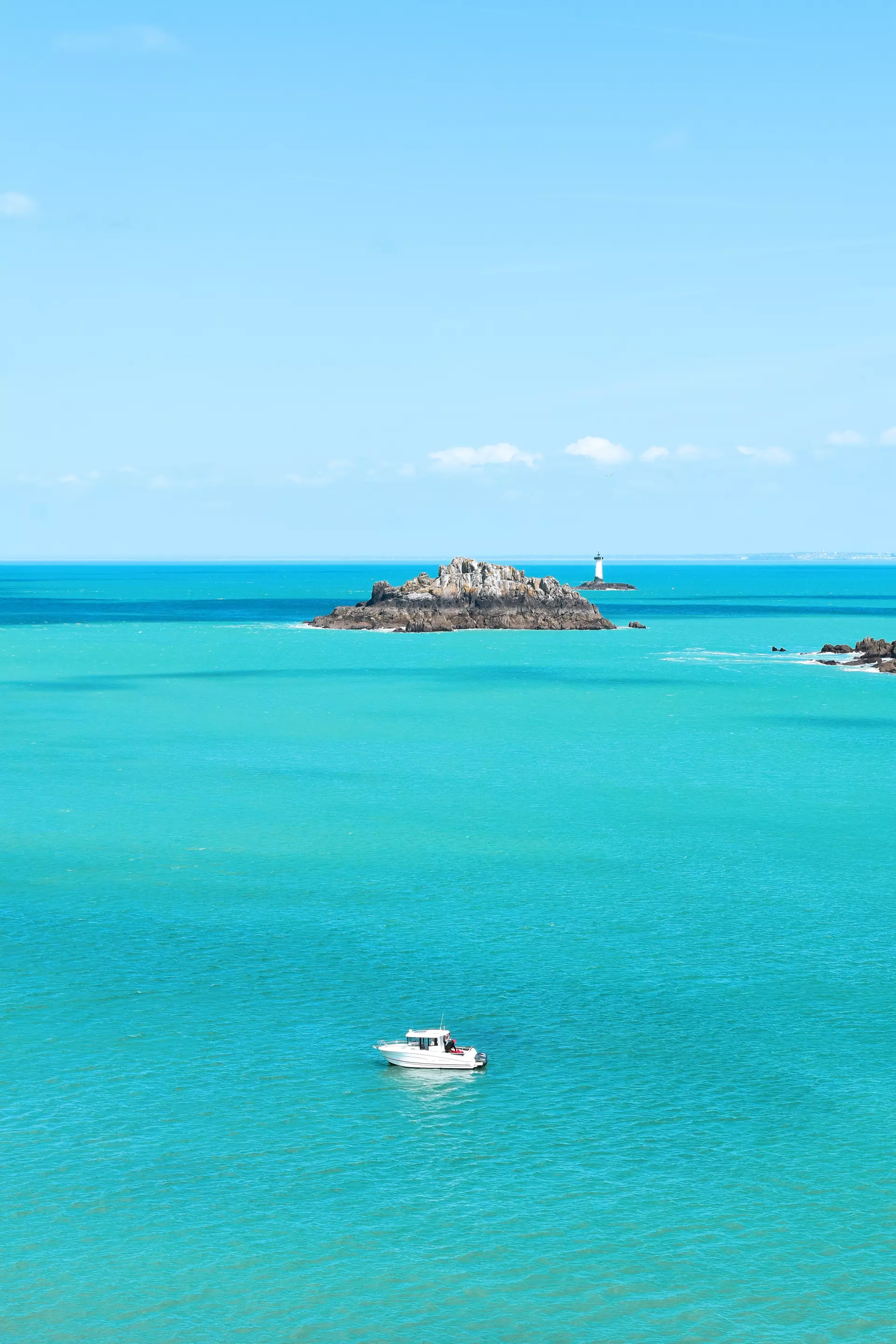 Kleines weißes Boot fährt auf türkisfarbenem Meer vor der Felseninsel mit Leuchtturm in der Bretagne bei Cancale