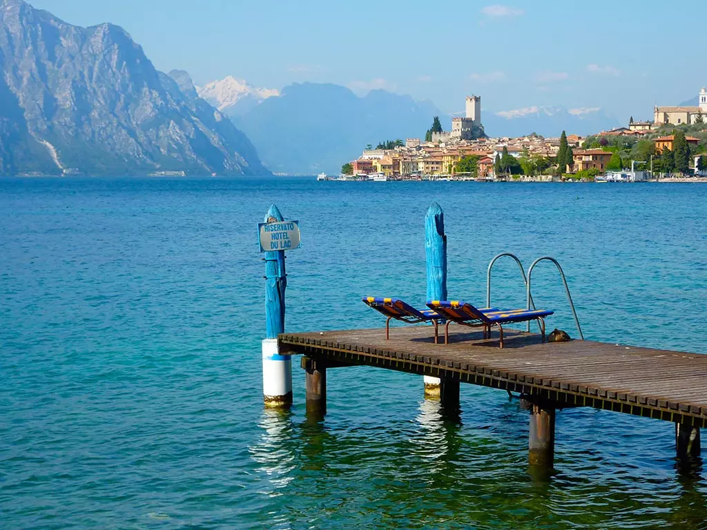 Holzsteg am Gardasee Holzsteg mit zwei Liegestühlen am Gardasee, im Hintergrund Berge und ein historisches Dorf mit Turm