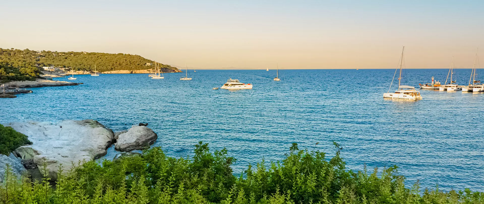 Küstenlandschaft in Griechenland mit grünem Buschwerk, Felsen im Vordergrund und mehreren Segel- sowie Motorbooten im ruhigen Meer bei Sonnenuntergang