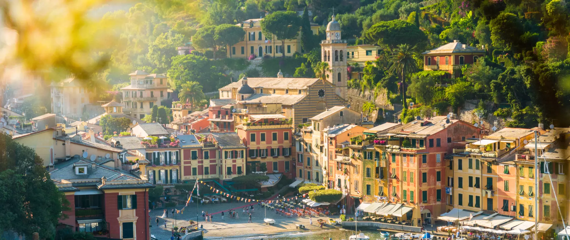 Bunte Häuser am Hafen eines italienischen Küstendorfs mit Sonnenschein und grünen Hügeln im Hintergrund