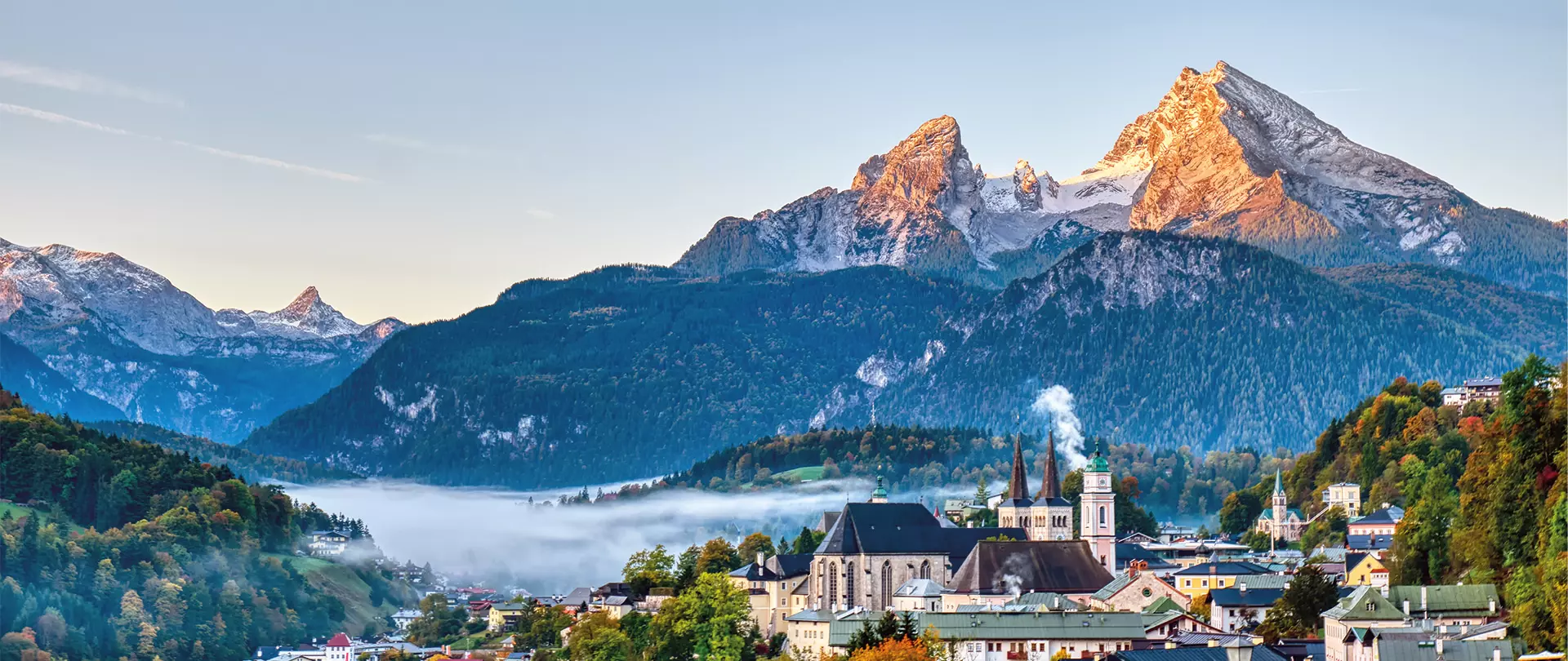 Bayerische Alpen mit Bergpanorama, nebligem Tal und traditionellem Dorf mit Kirche im Vordergrund bei klarem Himmel