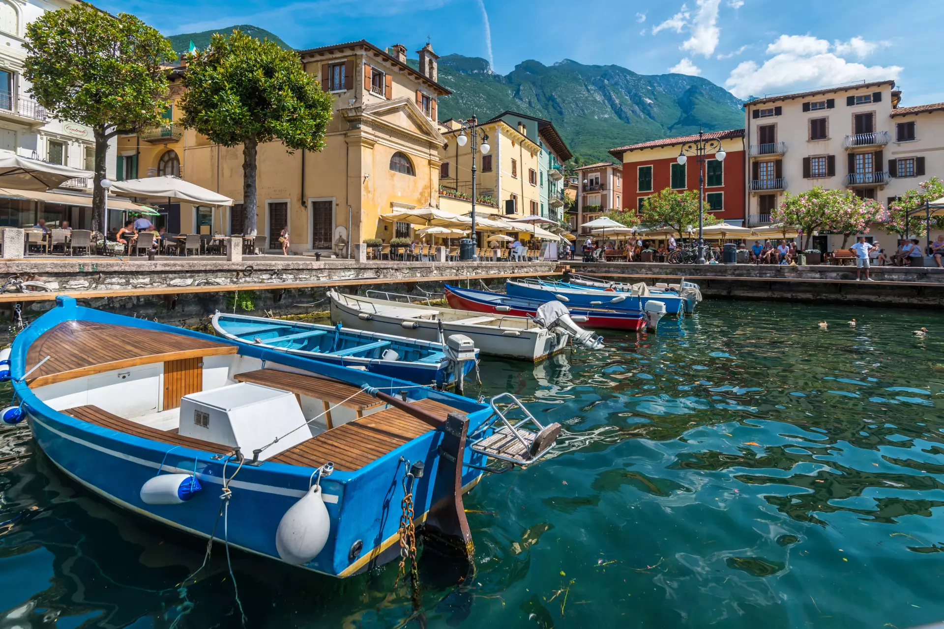 Boote am Ufer eines Hafenbeckens mit bunten Häusern und Bergen im Hintergrund am Gardasee