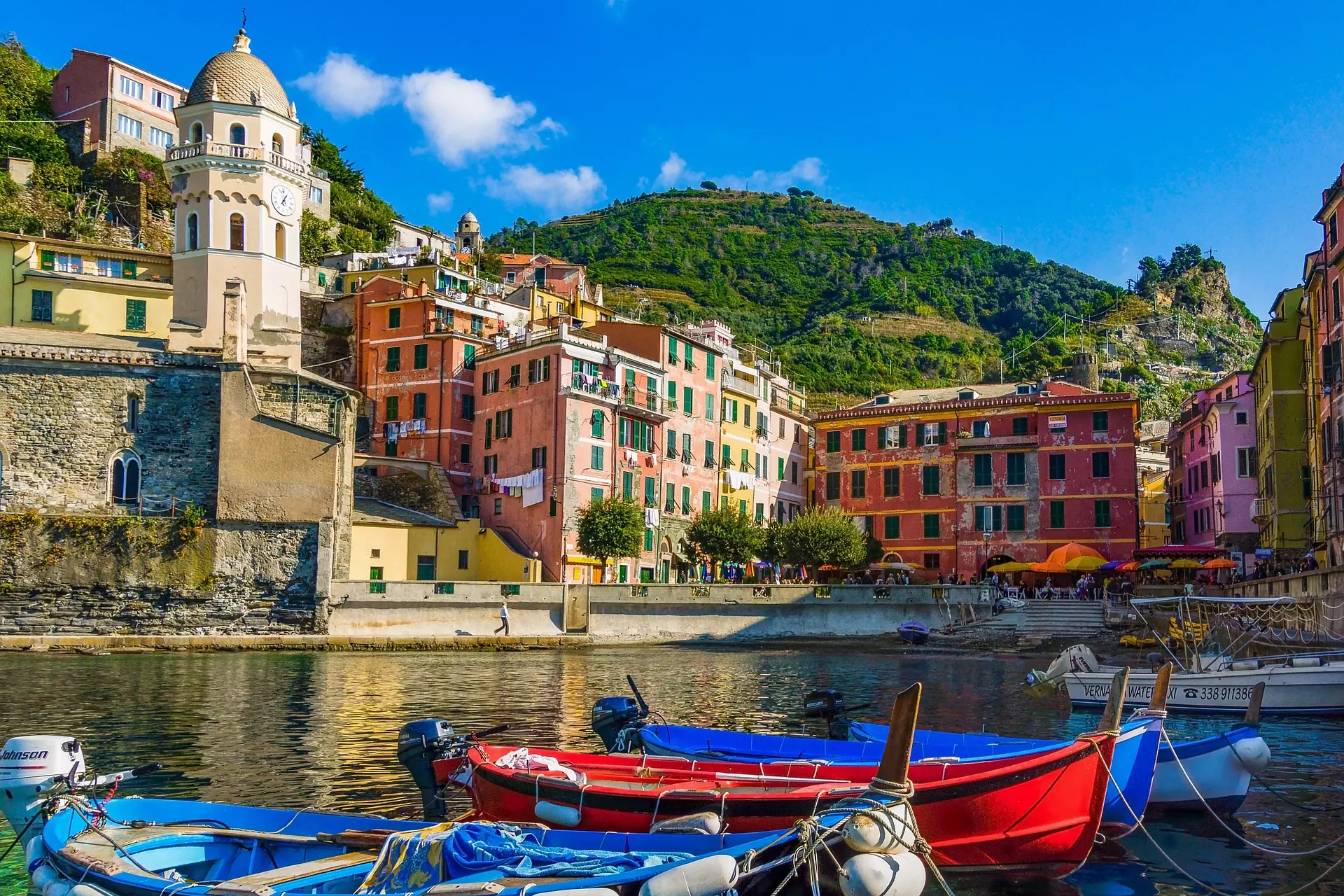 Bunte Boote im Wasser vor pastellfarbenen Häusern und einem Glockenturm in einem ligurischen Küstenort mit grünem Hügel im Hintergrund