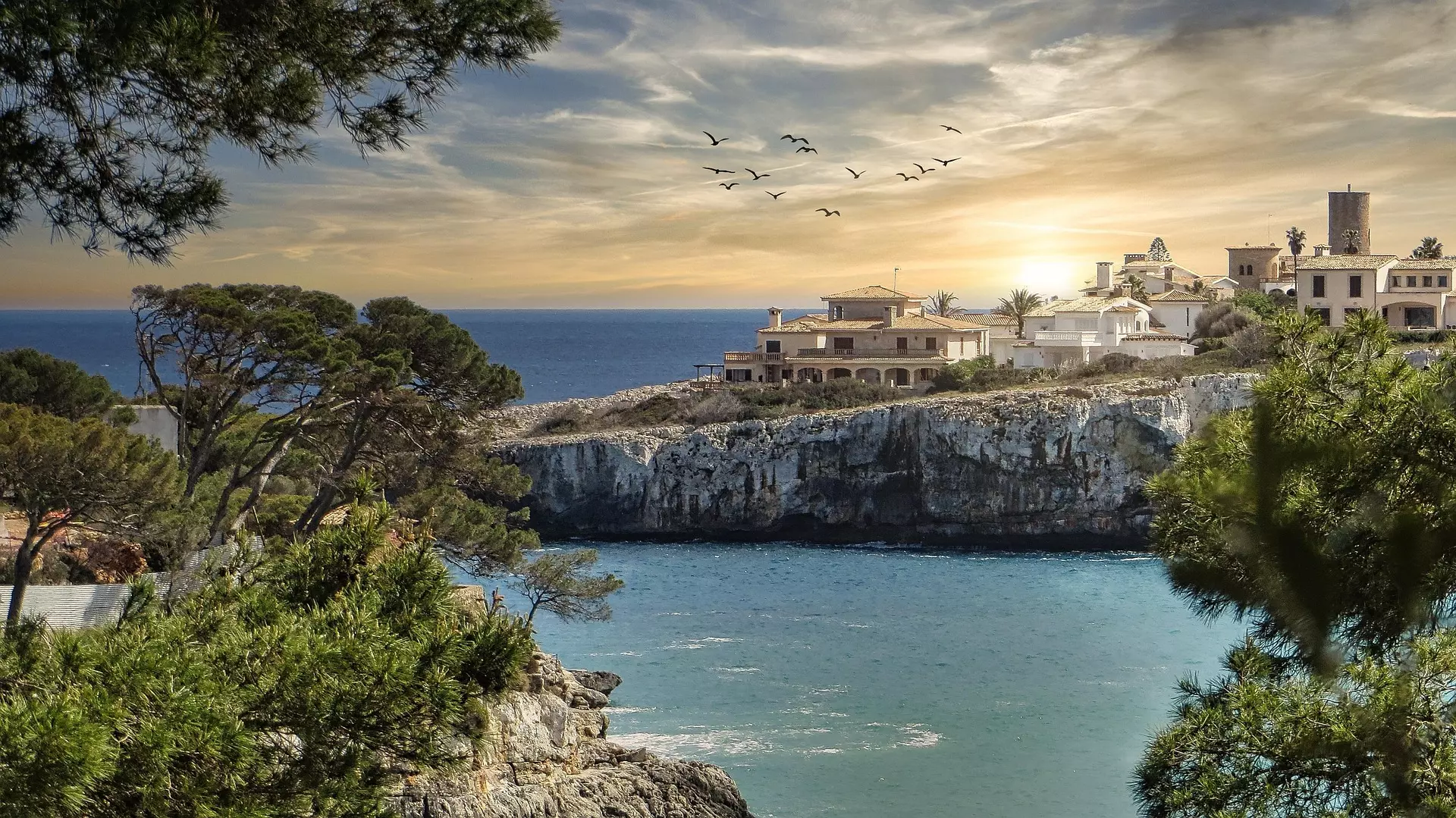 Küstenlandschaft Mallorcas Klippen- und Küstenlandschaft auf Mallorca mit Meer, Bäumen, Häusern auf dem Felsen und Vögeln im Sonnenuntergangshimmel