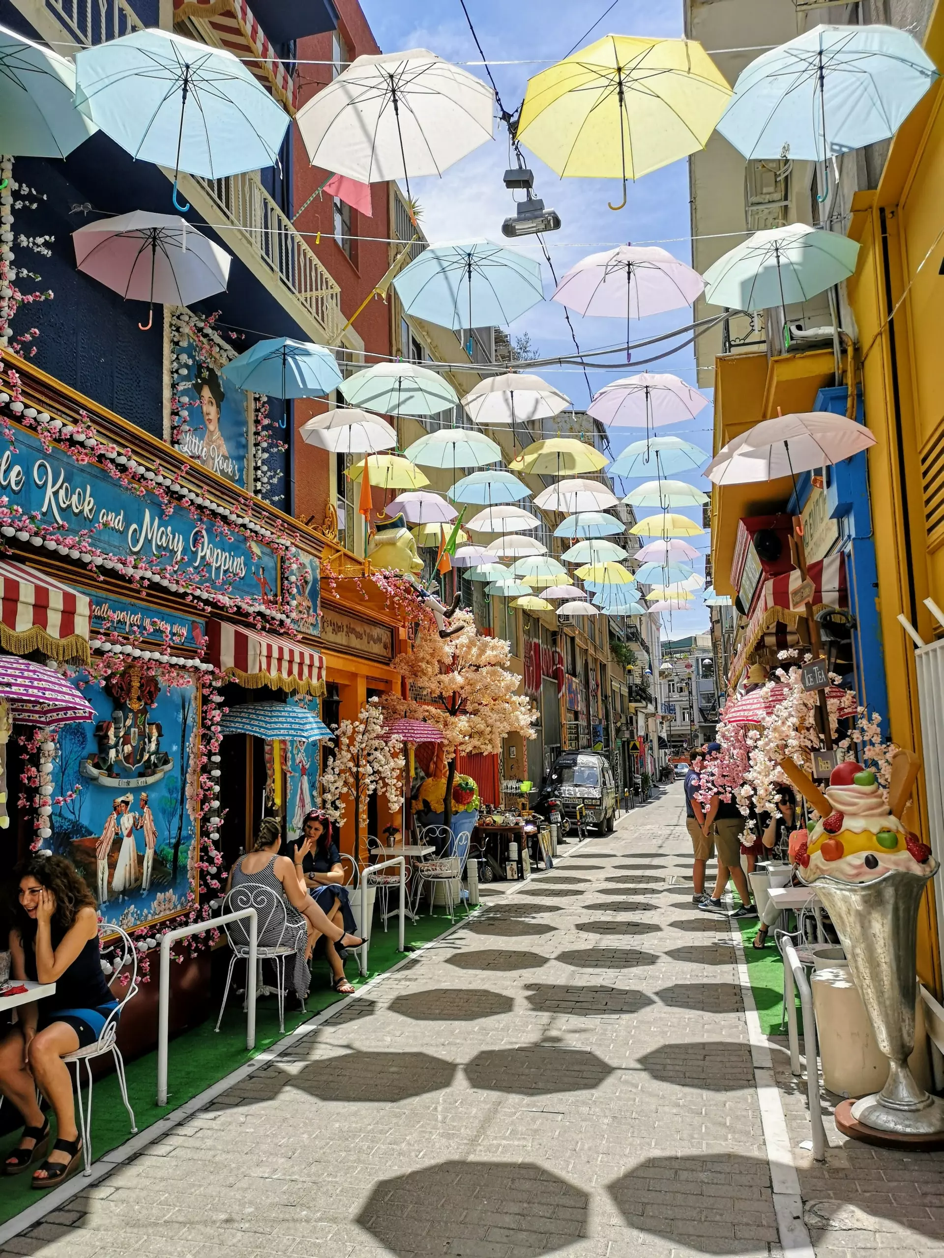 Bunte Regenschirme hängen über einer engen Straße in Athen mit Cafés, Sitzgelegenheiten und blühenden Dekorationen an den Gebäuden